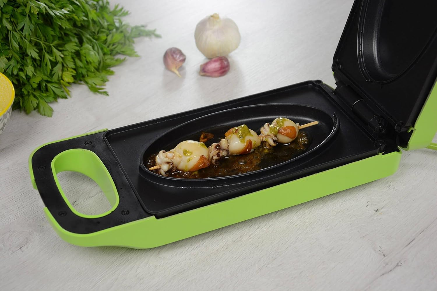 Griglia elettrica verde con spiedini di cibo all'interno. Aglio e prezzemolo sono sul tavolo accanto.