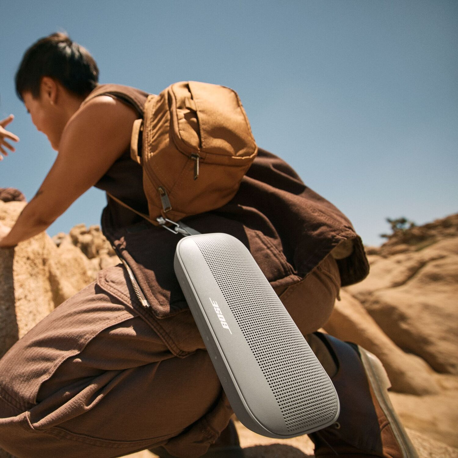 A person hiking with a grey Bose speaker attached to a brown backpack.