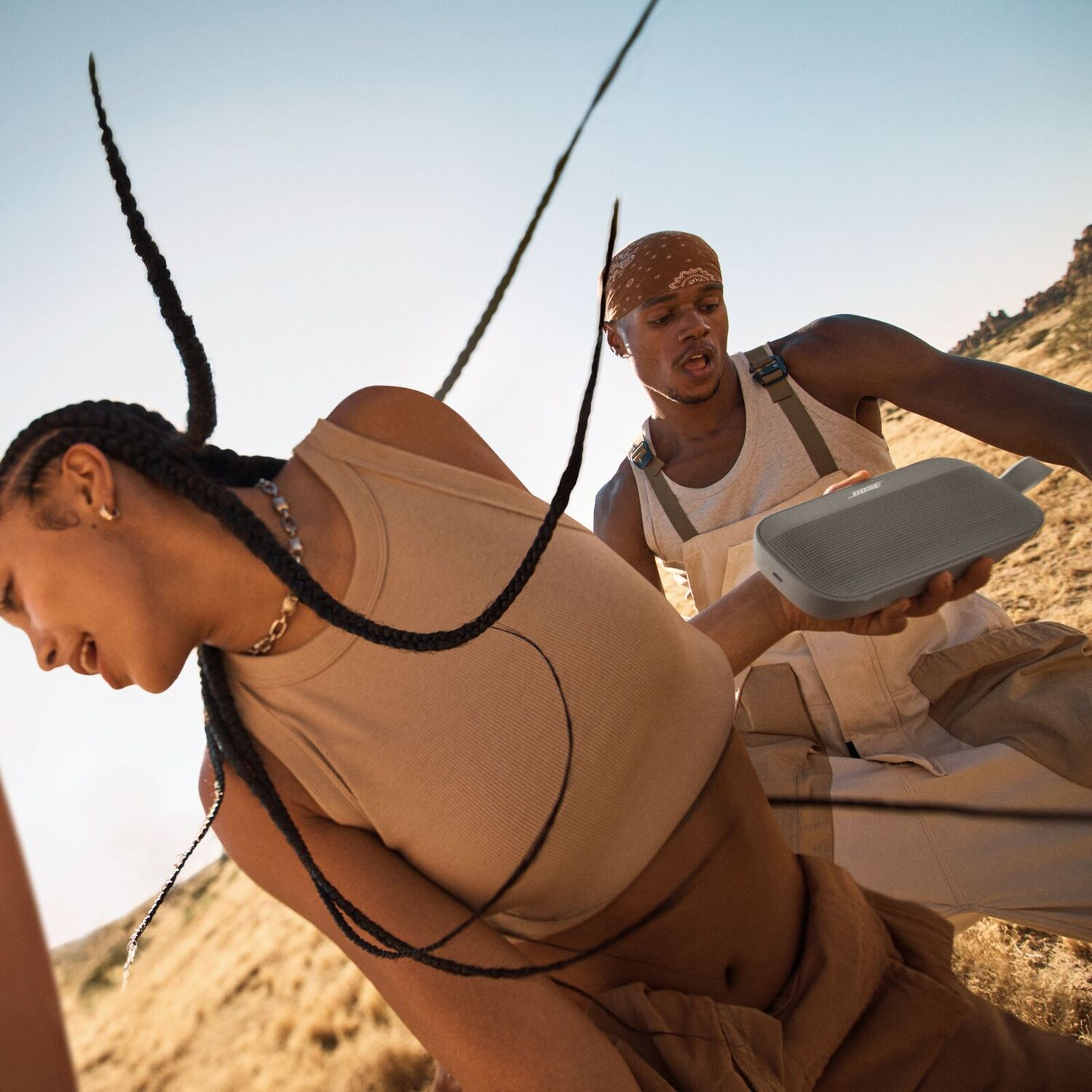 Two people interacting with a gray speaker in an outdoor setting with a desert backdrop.