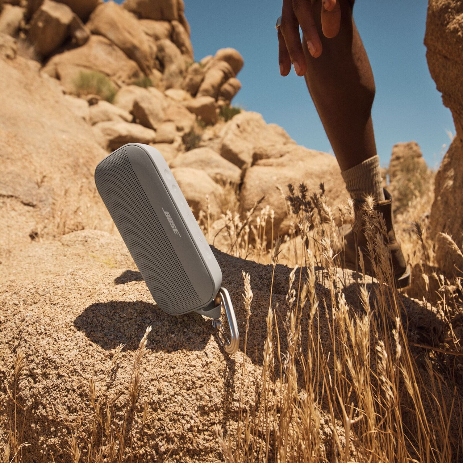 Gray Bose speaker on rock. Person's leg and hand visible. Desert background.