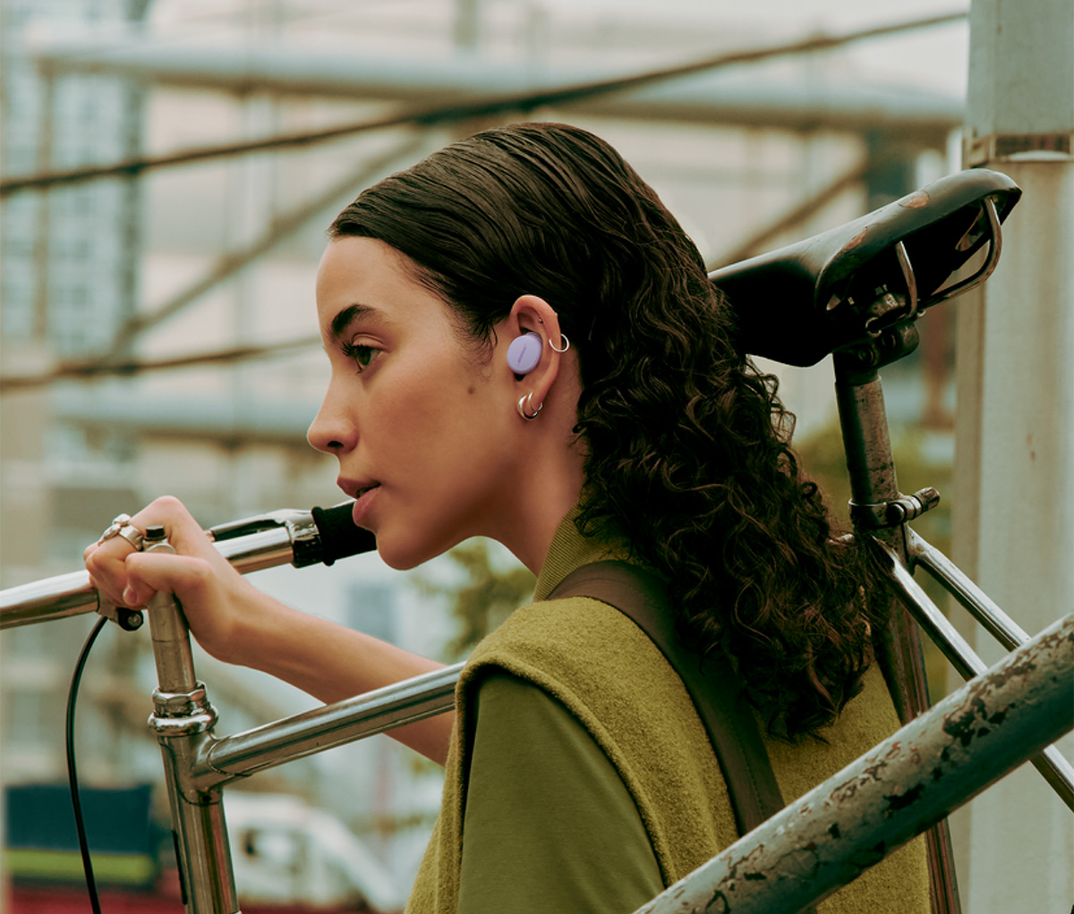Donna con auricolare viola nell'orecchio, che tiene una bicicletta. Indossa un gilet e una camicia verdi.