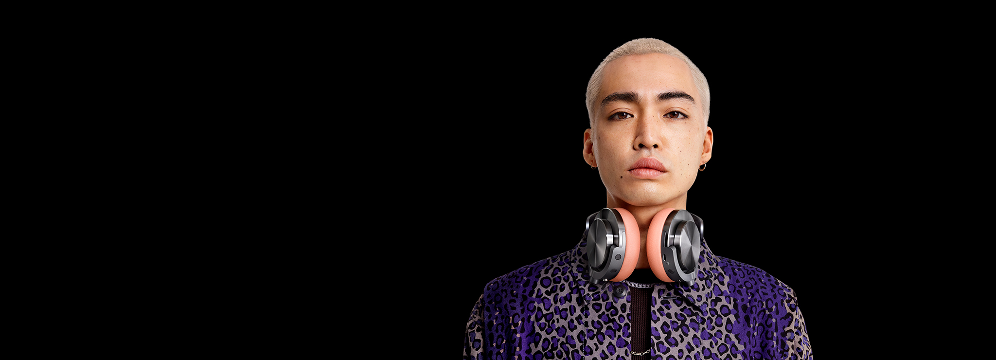 Una persona con cabello rubio y auriculares alrededor del cuello, sobre un fondo negro. Lleva una camisa estampada.