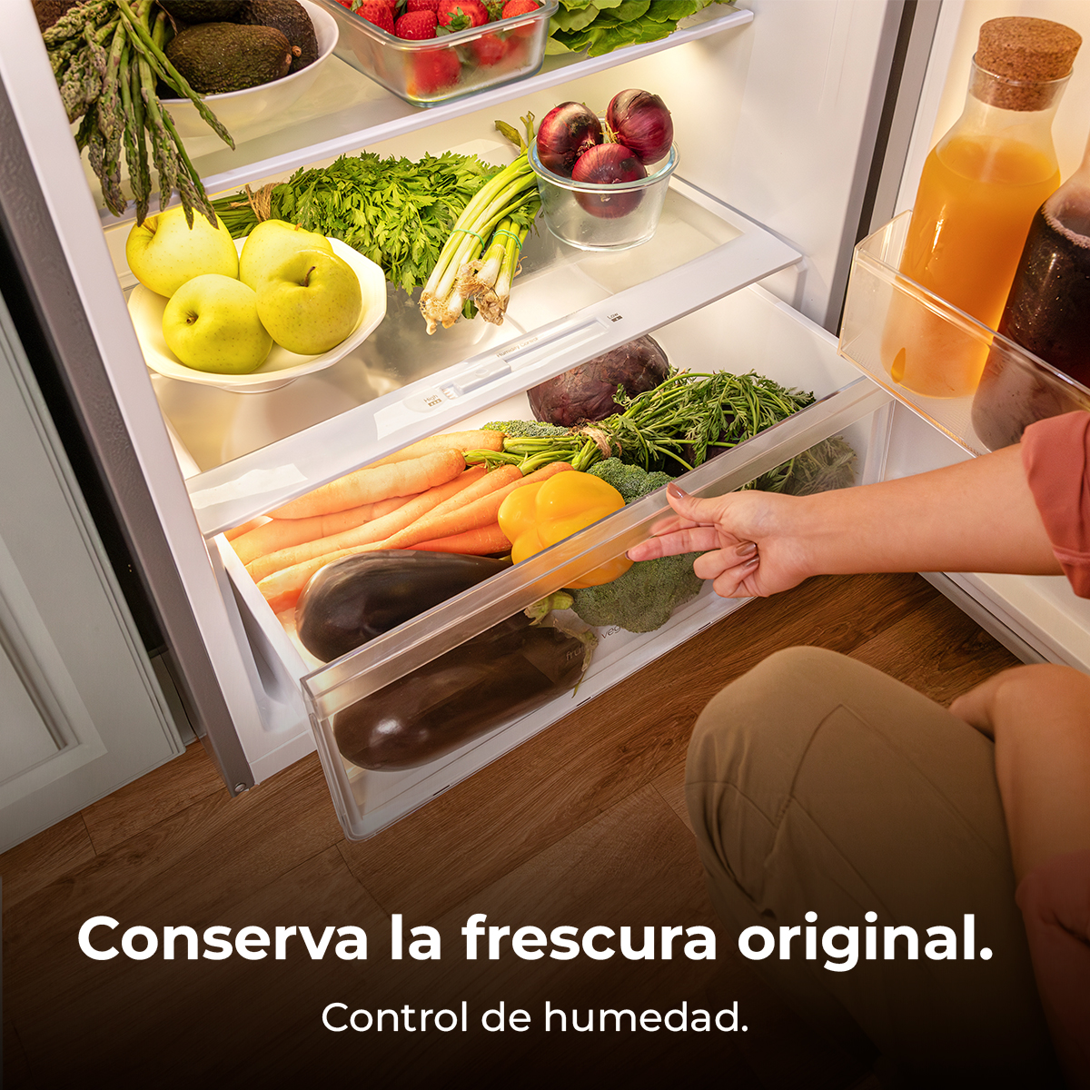 Refrigerador abierto con verduras y frutas en cajones, la mano de una persona se acerca.