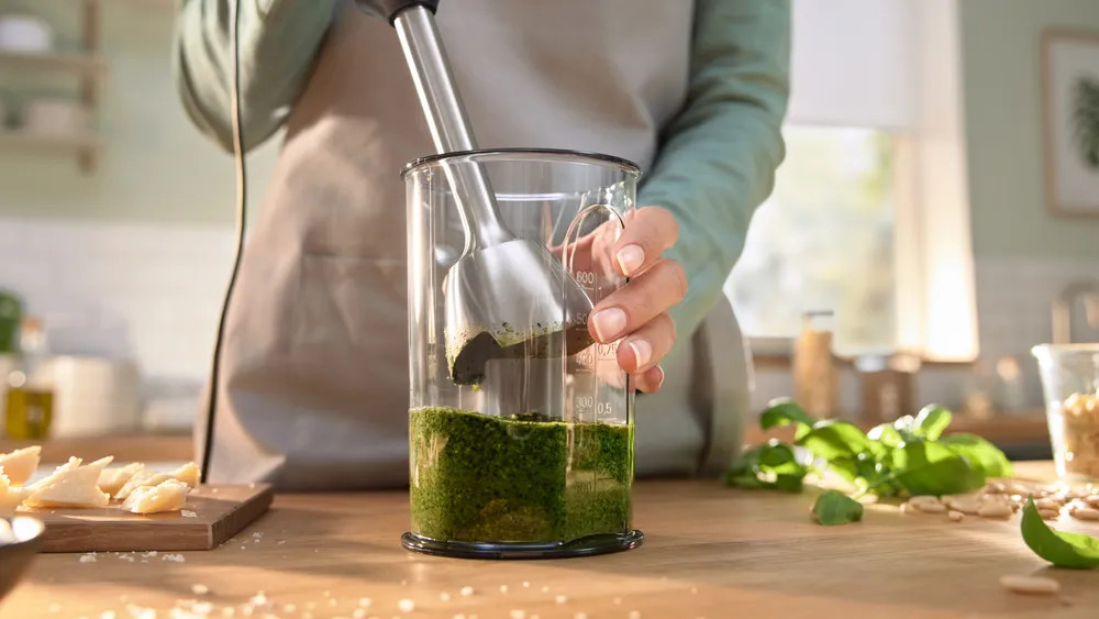 Osoba miksująca pesto w szklanym słoiku blenderem ręcznym, składniki na stole.