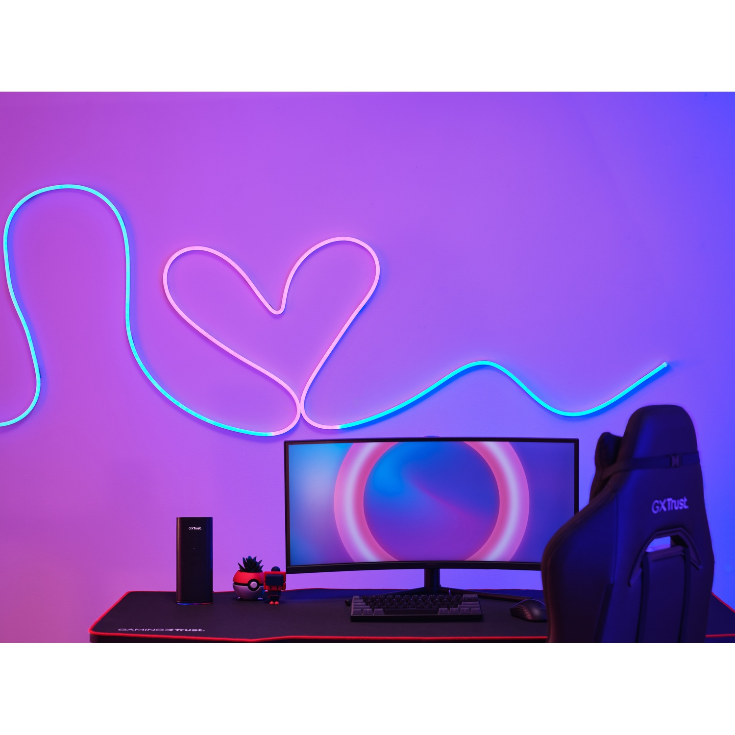 Un'impostazione di gioco con monitor, tastiera e un cuore al neon su un muro viola. È presente un altoparlante nero.