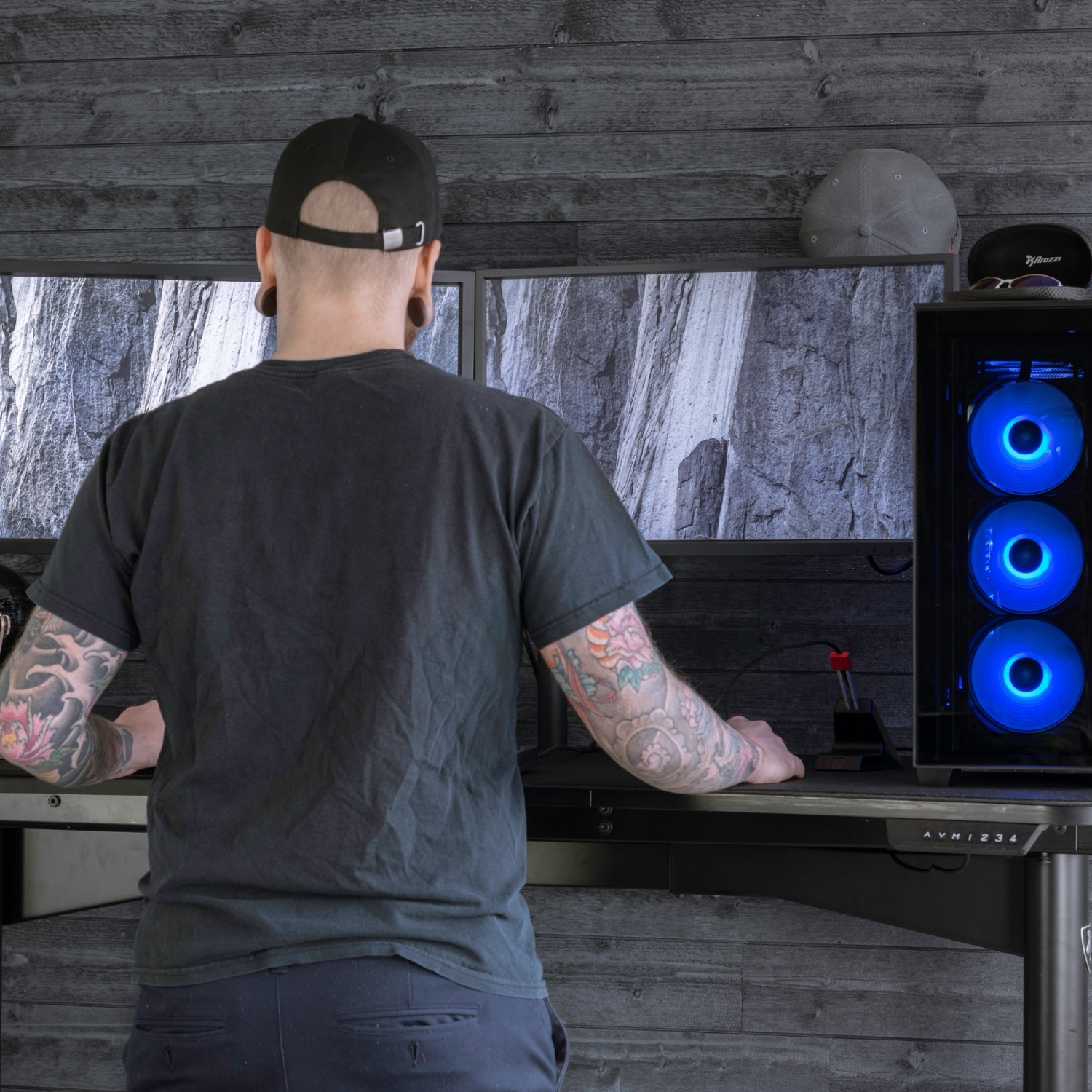 Mann am Schreibtisch mit zwei Monitoren, Gaming-PC mit blauen Lichtern, schwarzer Hut.