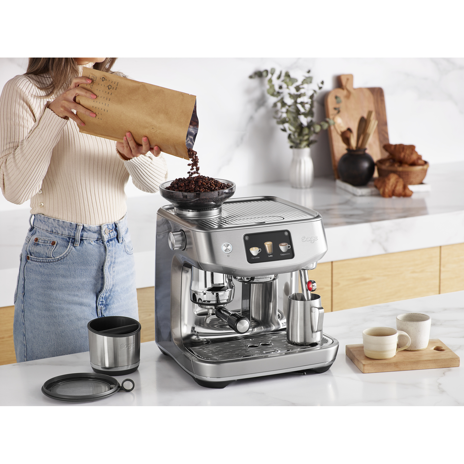 Femme versant des grains de café dans une machine à expresso Sage sur un comptoir blanc.