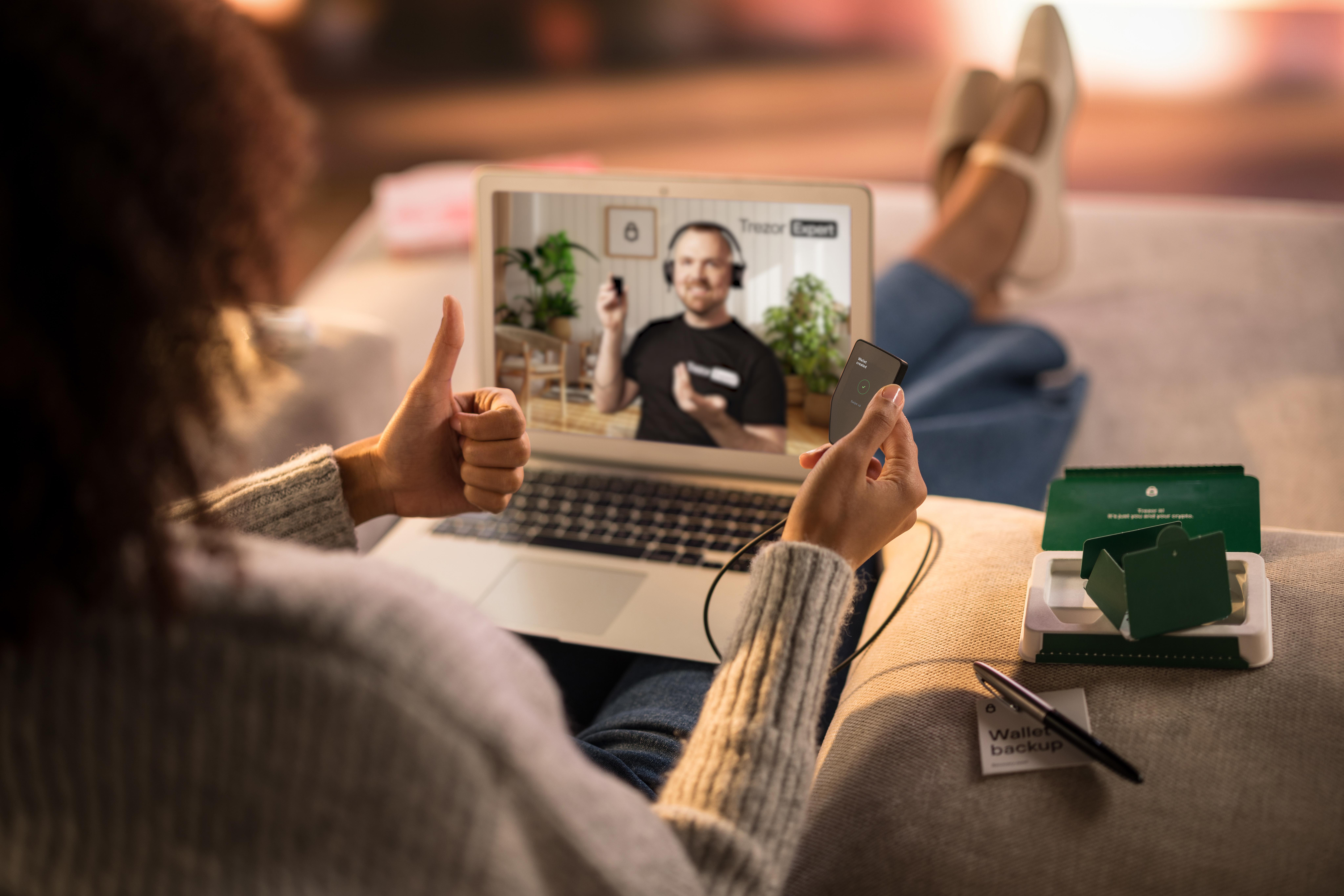 Frau gibt Daumen hoch, schaut Videoanruf auf Laptop. Trezor Hardware-Wallet sichtbar.