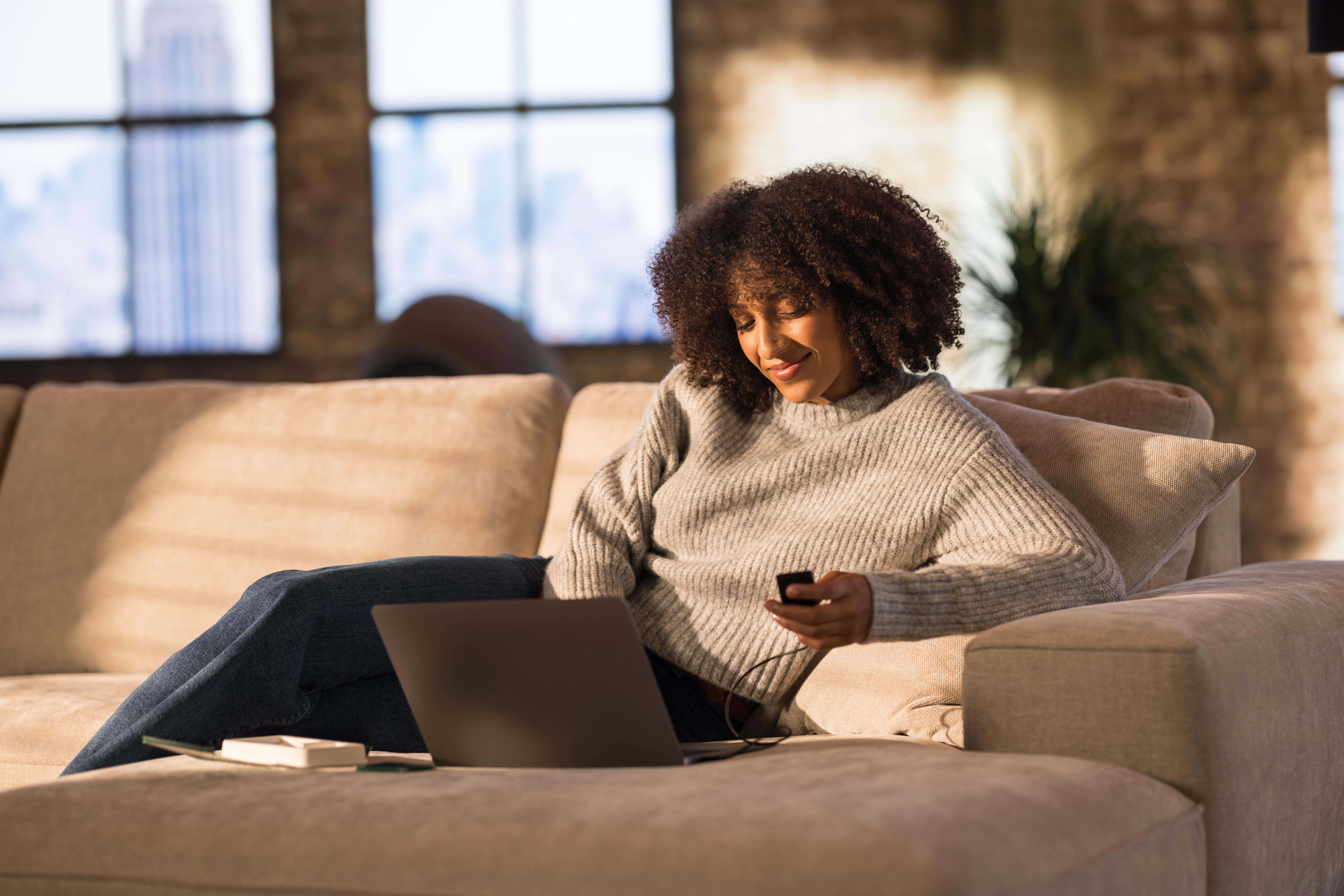 Eine Frau benutzt einen Laptop und ein Telefon, während sie auf einer Couch sitzt.