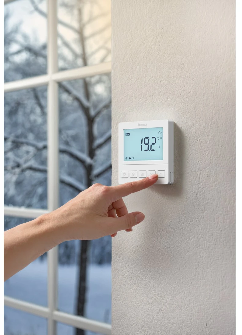 Eine Hand drückt eine Taste an einem weißen Thermostat an einer strukturierten Wand in Fensternähe.