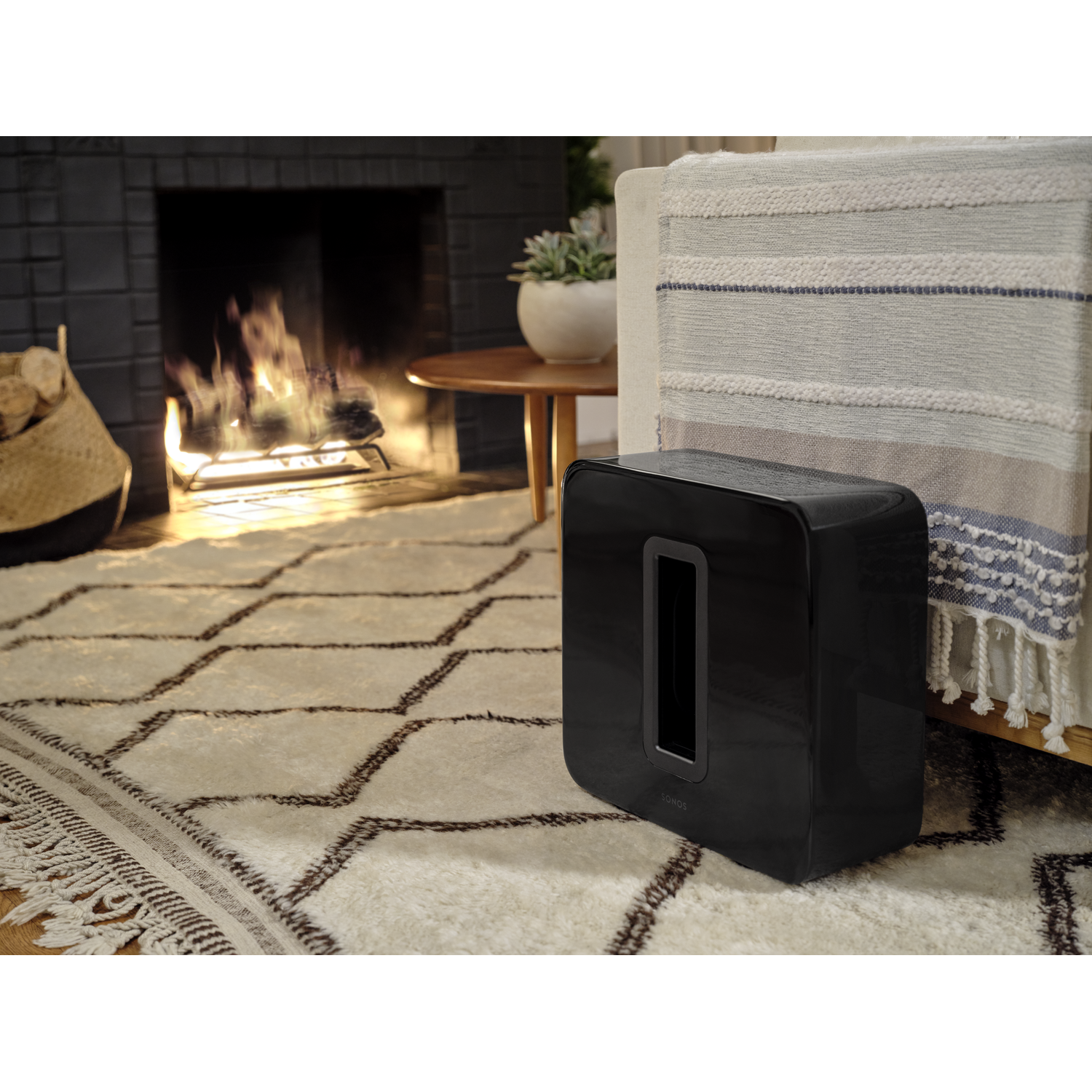 Zwarte Sonos subwoofer naast een bank, open haard op de achtergrond.