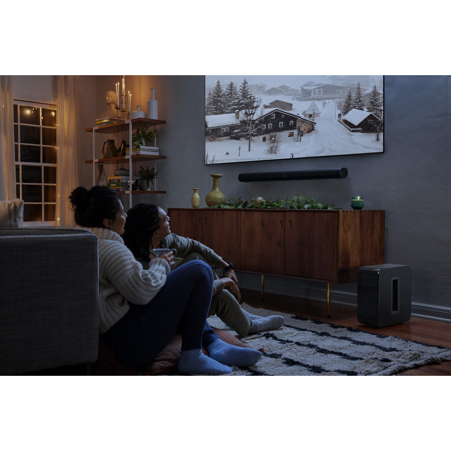 Twee mensen kijken tv met een besneeuwd landschap. Soundbar, subwoofer en kaarsen zijn aanwezig.