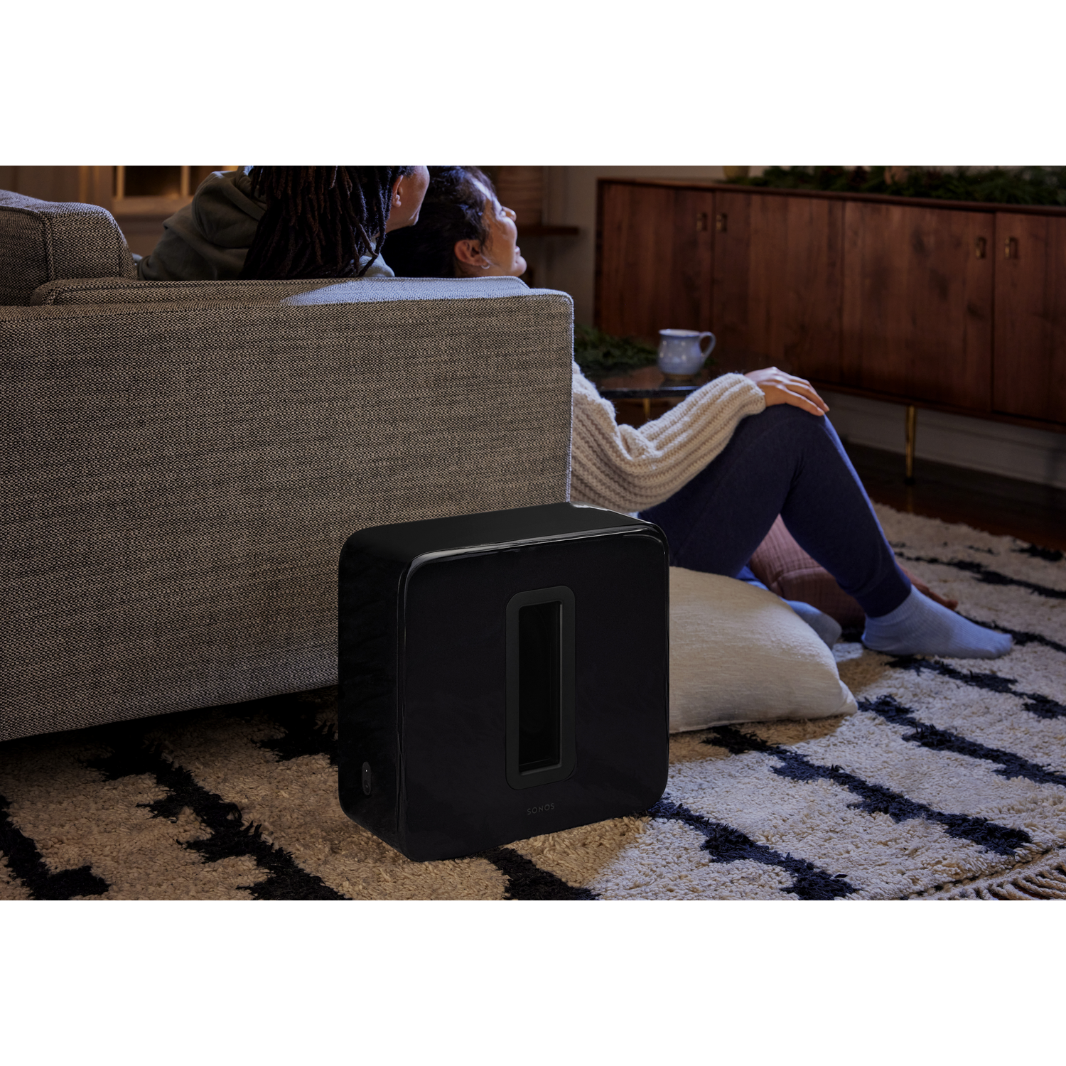 Zwarte subwoofer naast een bank. Twee personen op de achtergrond.