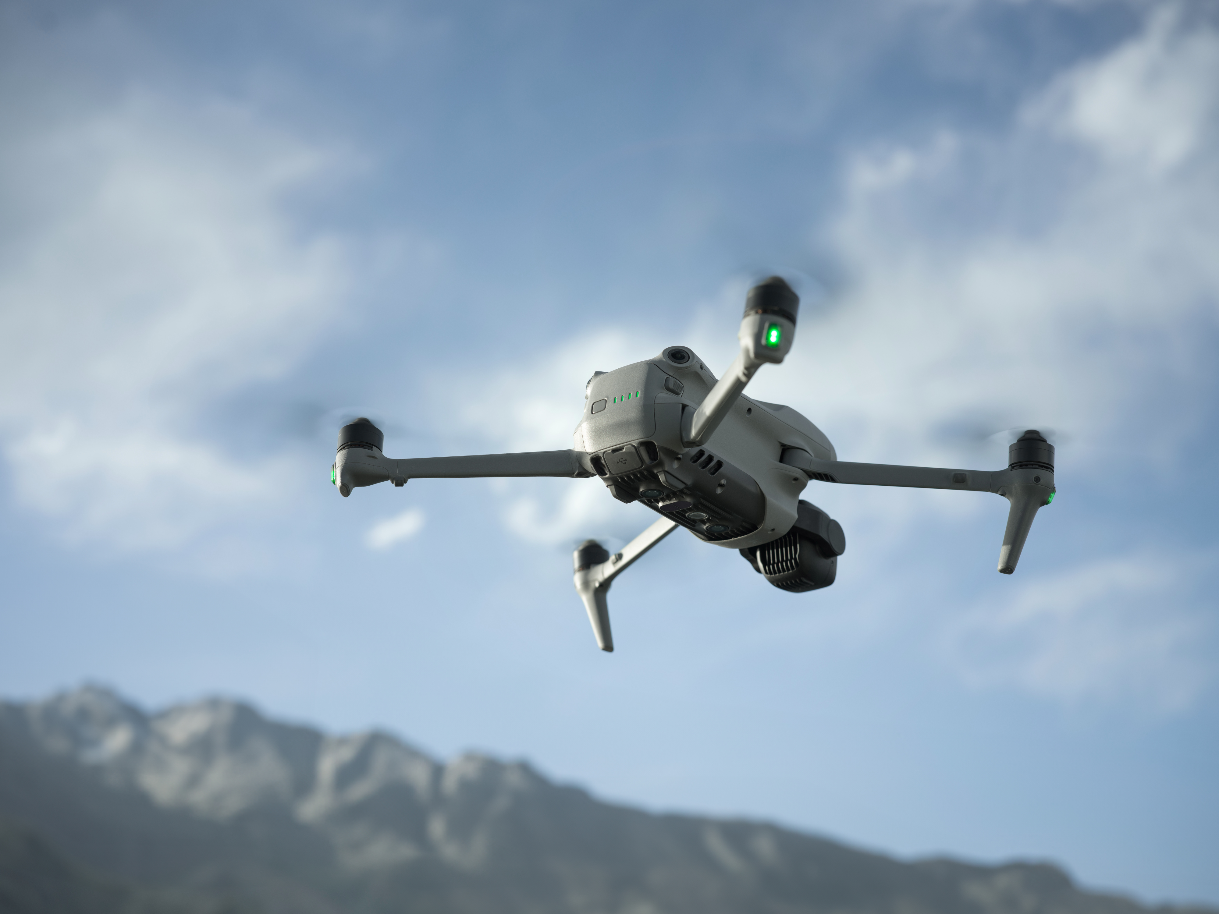 Un drone grigio vola contro un cielo blu con montagne sullo sfondo.