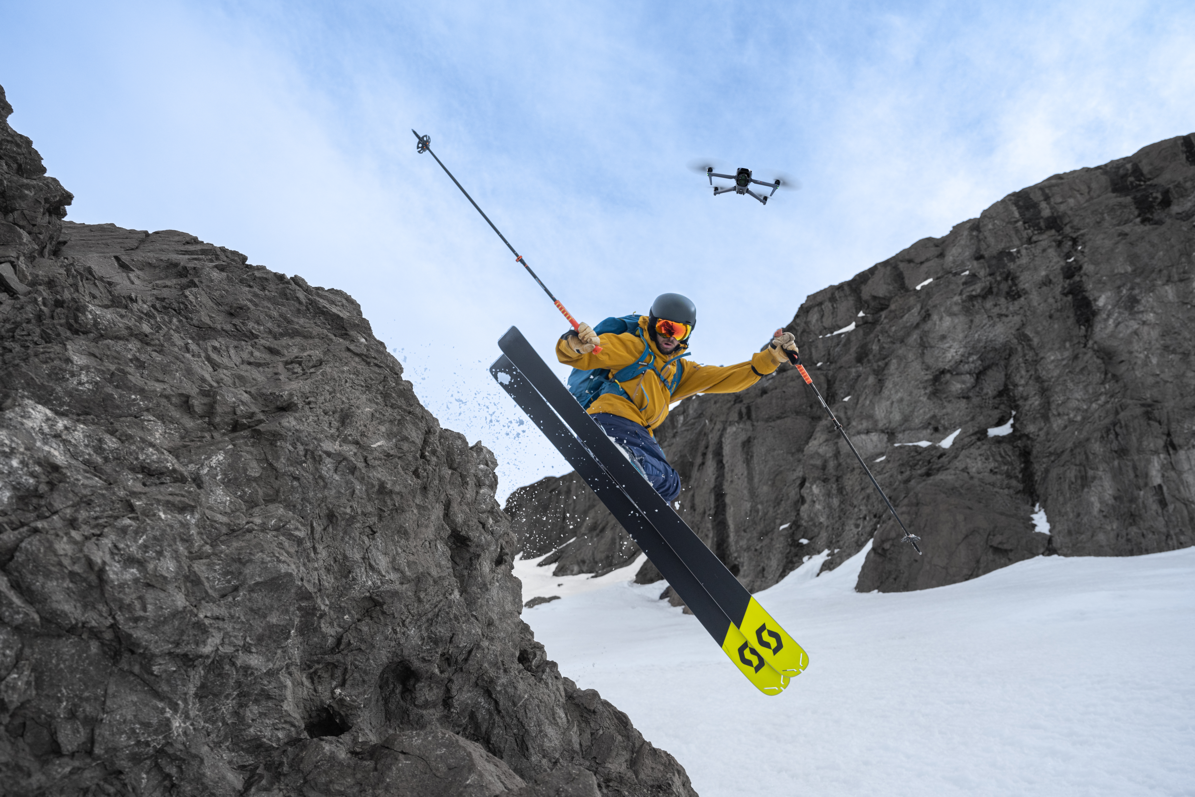 Scialpinista in aria sopra roccia. Un drone vola sopra. Lo sciatore indossa equipaggiamento giallo e nero.