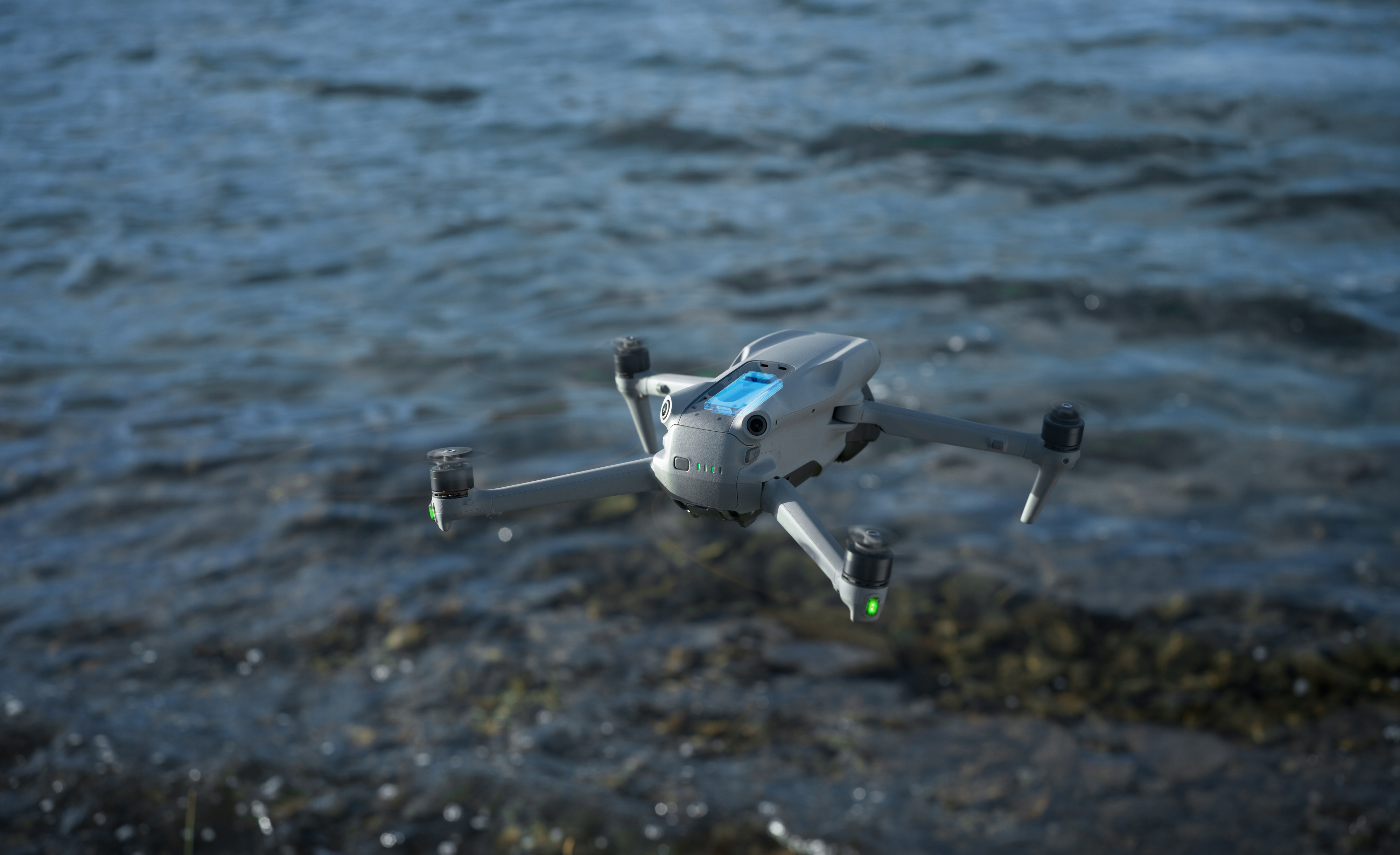 Un drone grigio è sospeso sull'acqua. Le eliche girano e il drone è a mezz'aria.