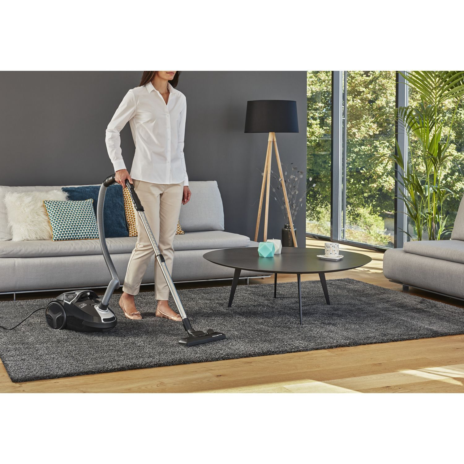 A woman vacuums a grey rug in a living room. There is a sofa, coffee table, and lamp.