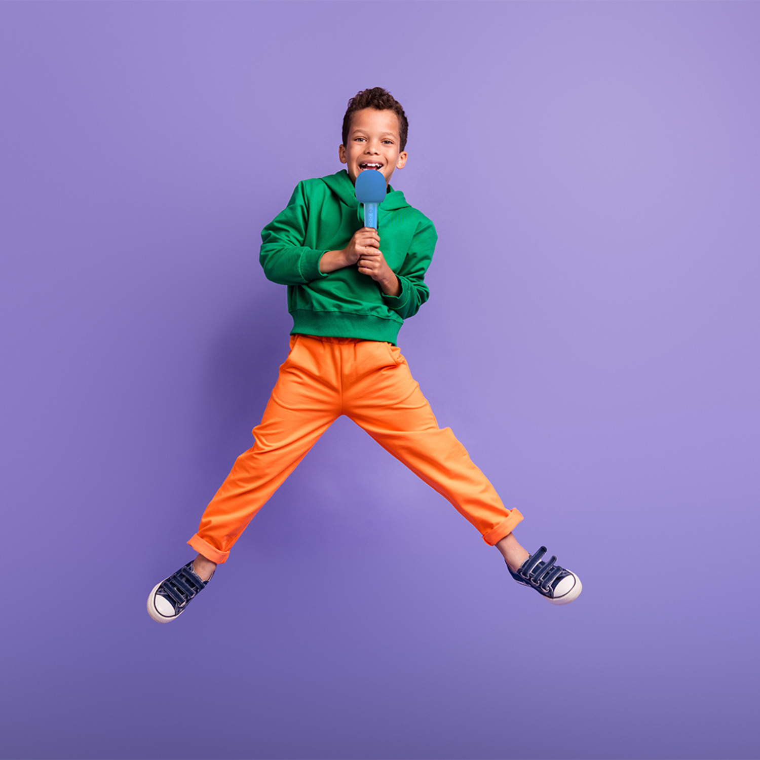 Un garçon en sweat à capuche vert et pantalon orange saute, tenant un micro bleu.