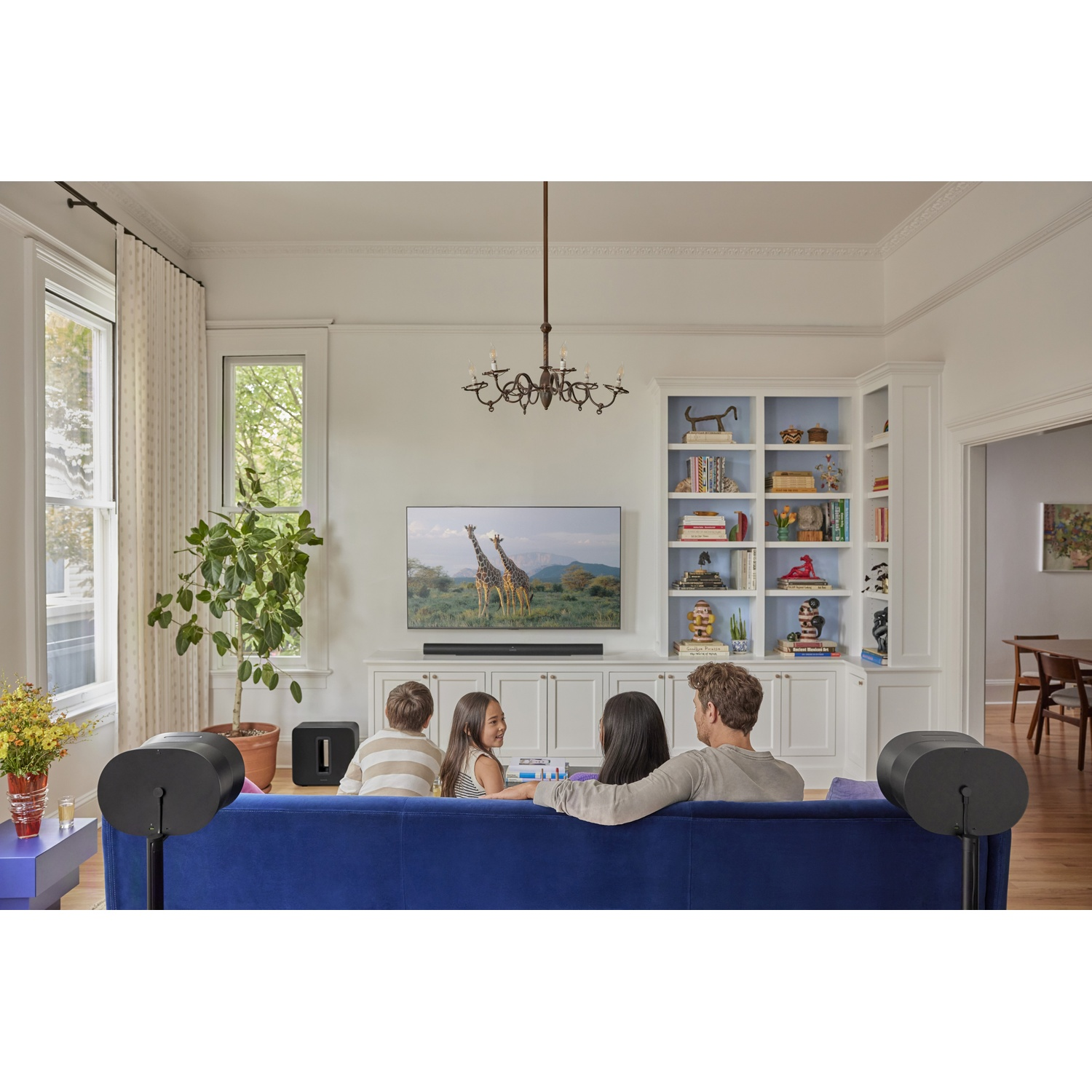 Famiglia guarda la TV su un divano blu. Un grande albero vicino alla finestra.