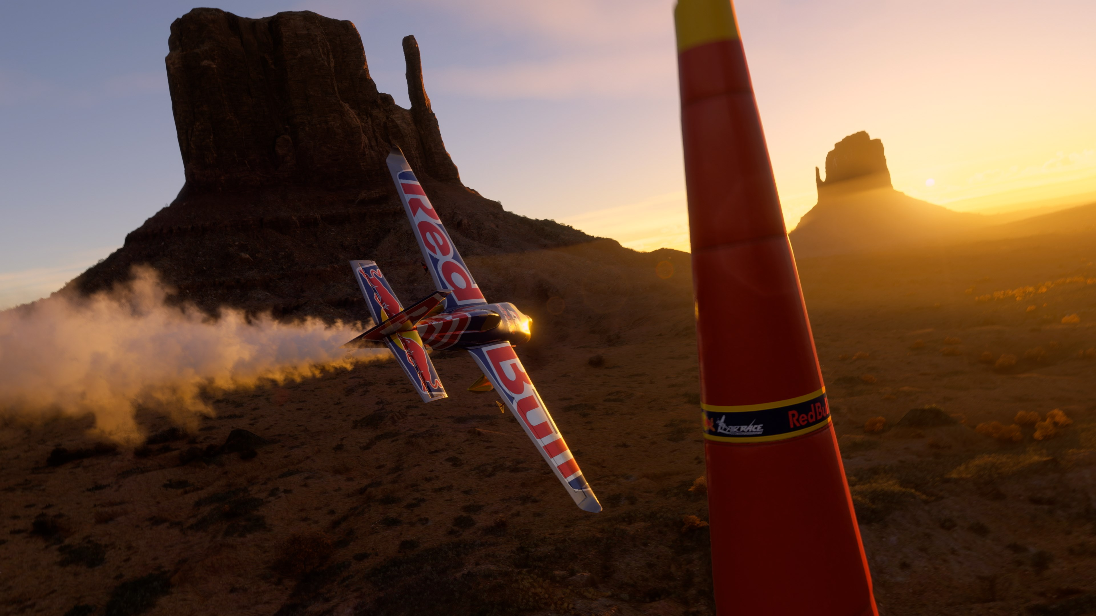 Red Bull Flugzeug im Flug in der Nähe einer Felsformation bei Sonnenuntergang. Ein roter Pylon im Vordergrund.