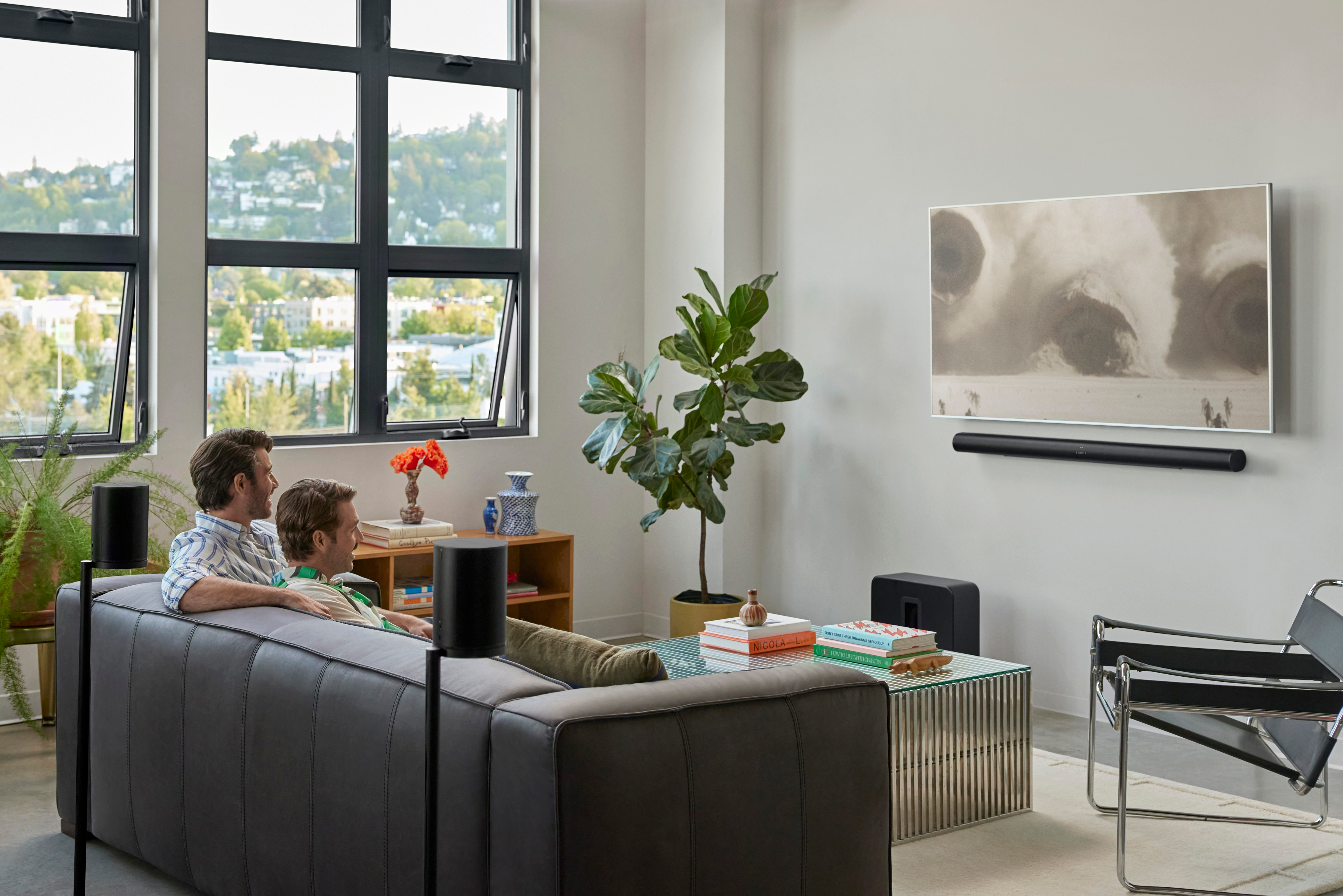 Deux hommes sur un canapé, une télévision avec une image au-dessus d'une barre de son et des enceintes.