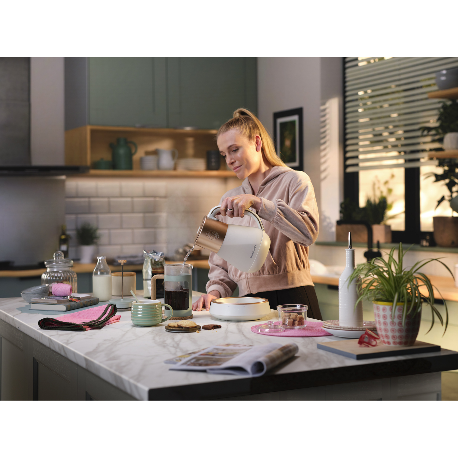Woman pouring water from a white and gold kettle into a glass. Kitchen counter with various items.