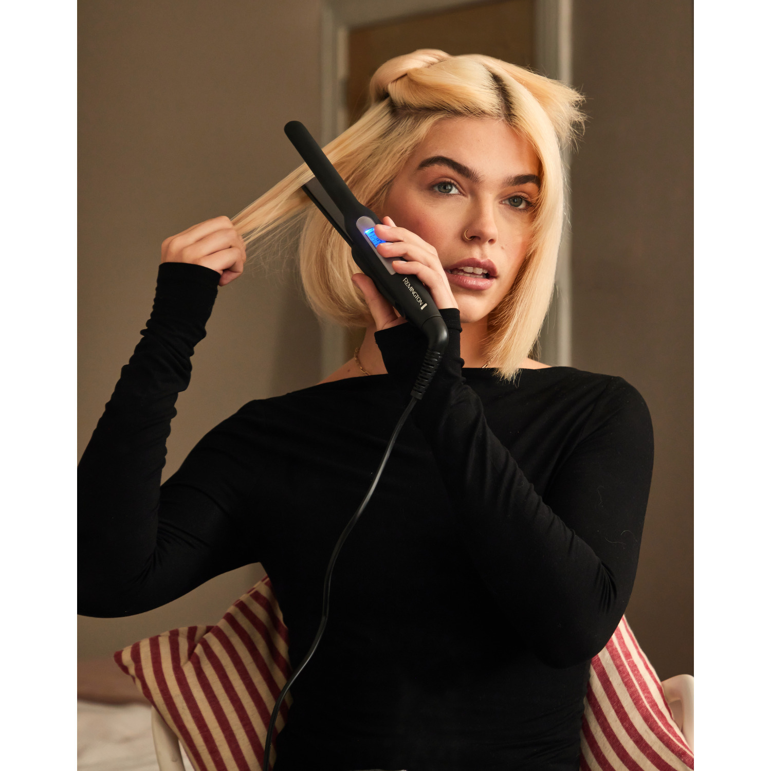Femme blonde avec un lisseur à la main, regardant l'appareil. Elle est dans une pièce.