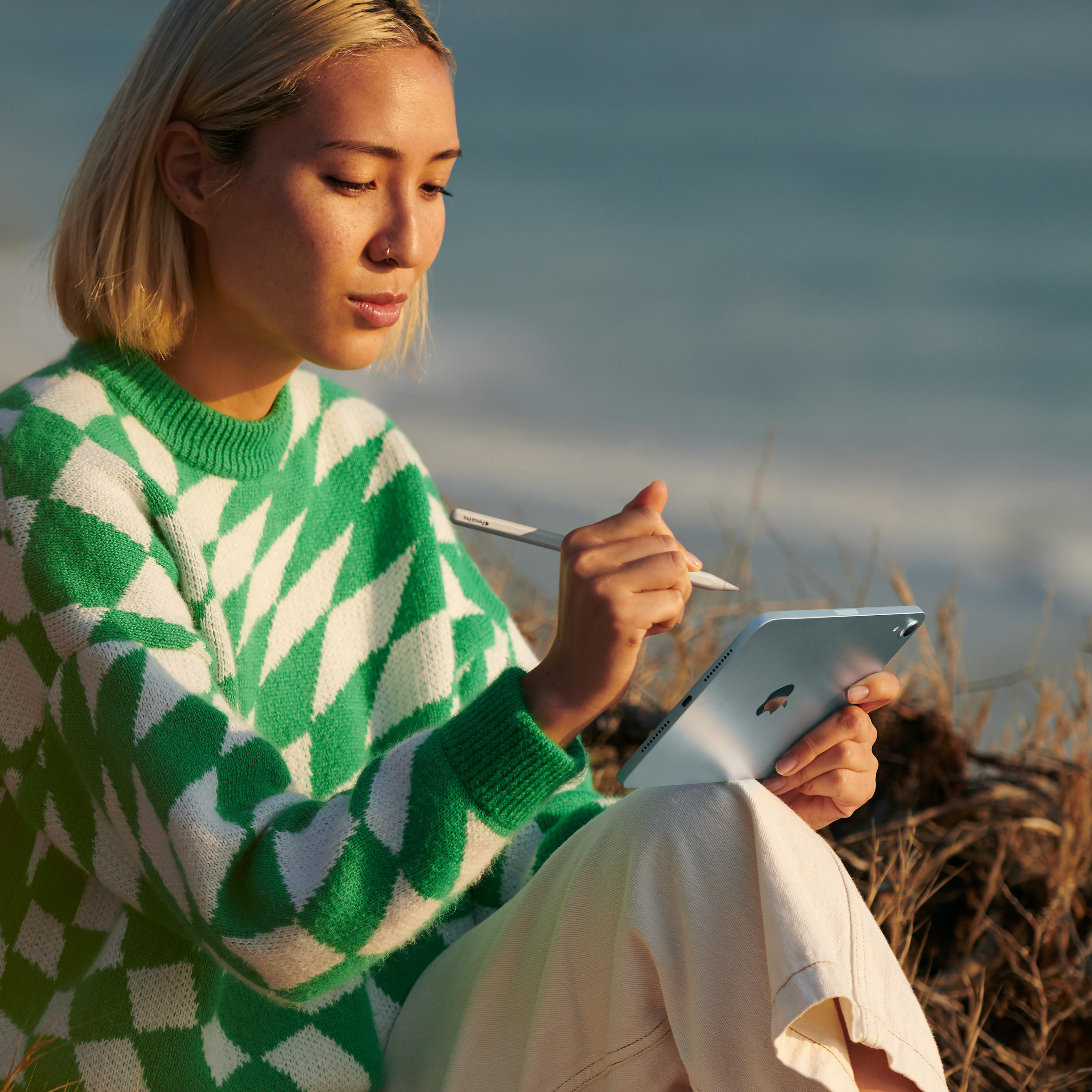 Frau benutzt ein iPad und einen Stift im Freien, sitzt im Gras in der Nähe des Wassers.
