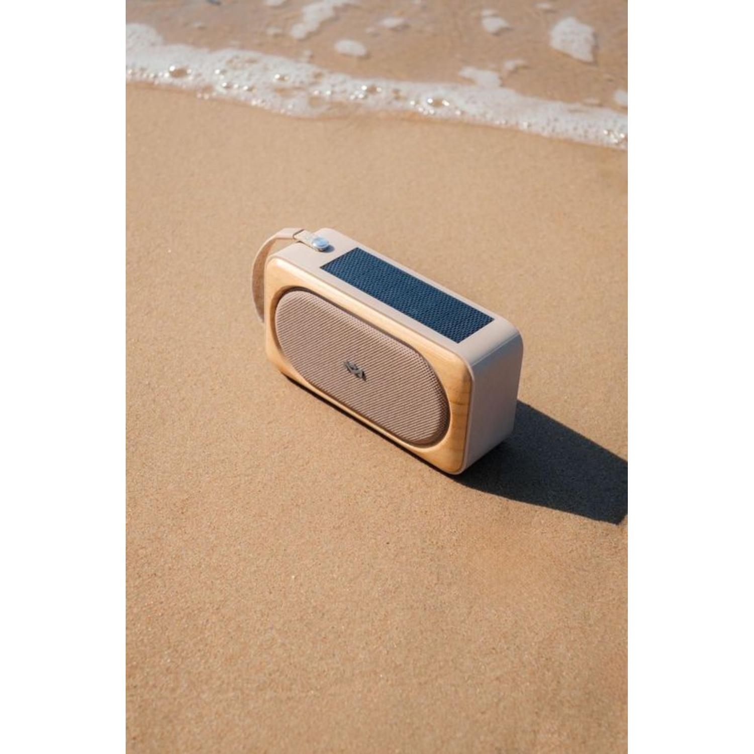 Een luidspreker, lichtbeige met houten accenten, op een zandstrand, bij de waterlijn.