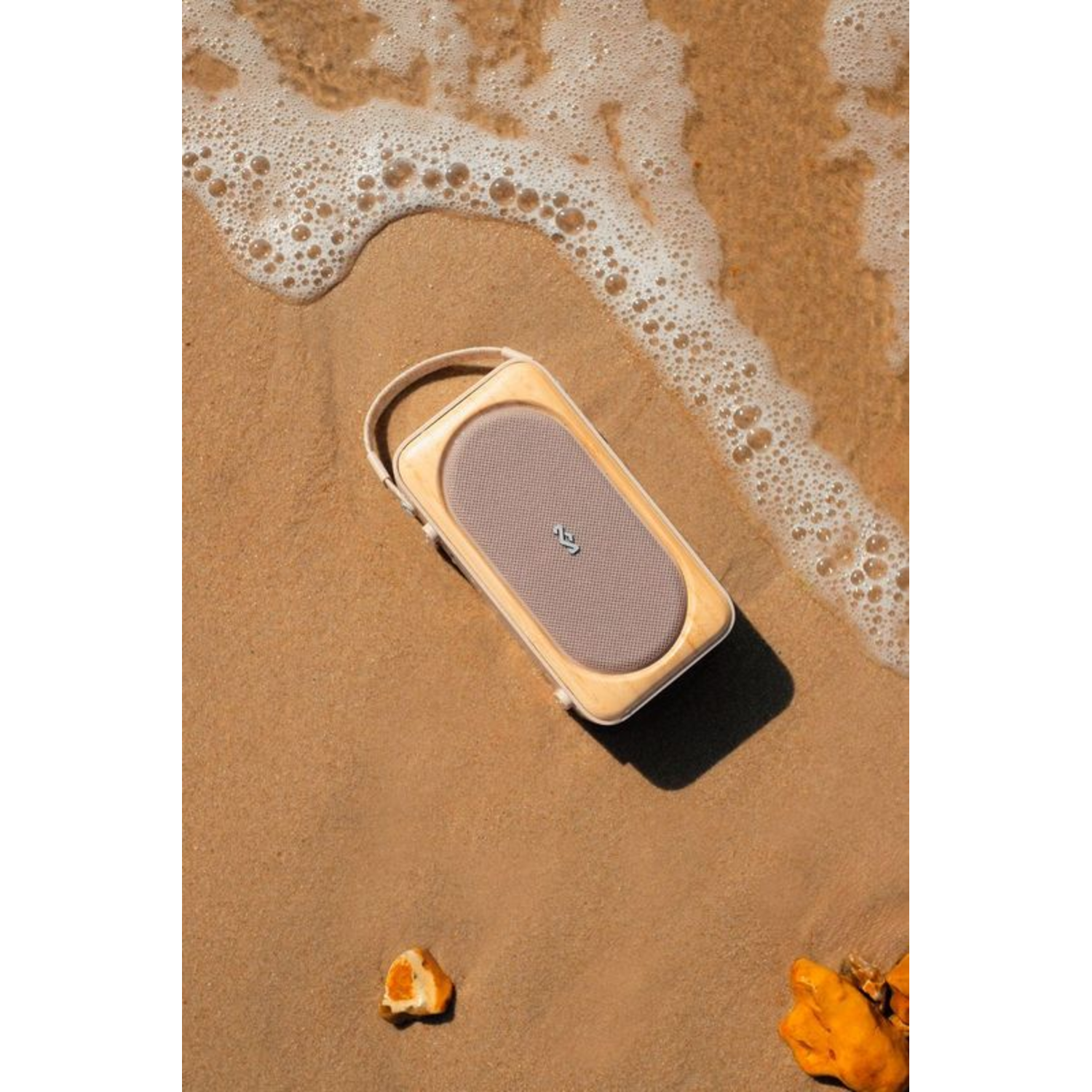 Een speaker op het strand met zand en golven. De speaker heeft een beige behuizing en een mesh voorkant.