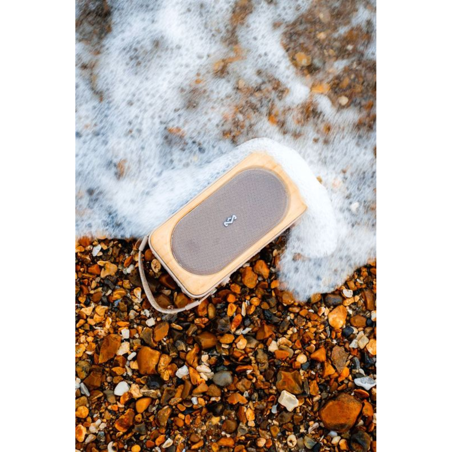 Een luidspreker op een strand met schuimig water. De luidspreker is lichtbruin en rechthoekig. Kiezels omringen hem.