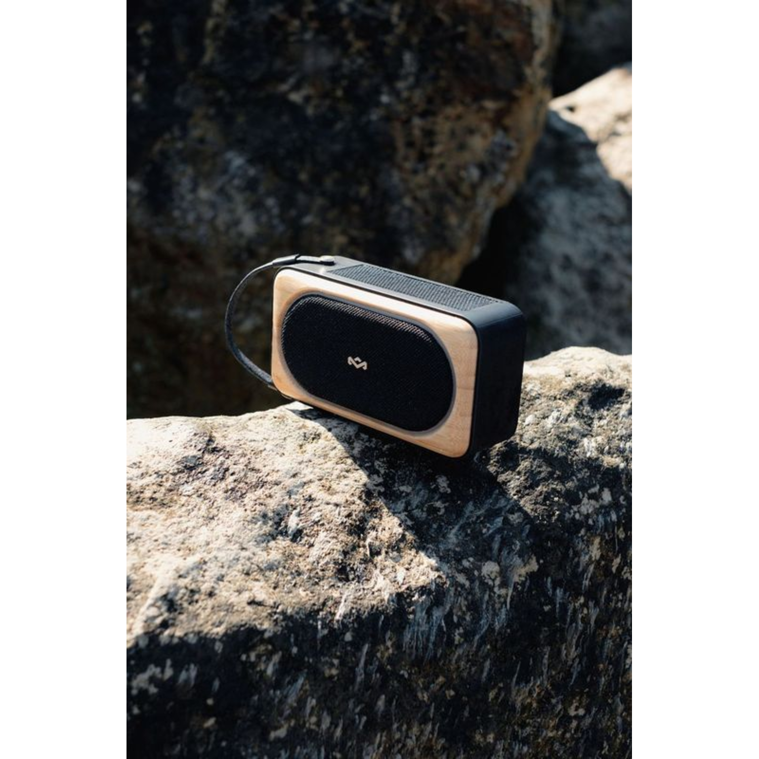 A black and wooden speaker sits on a rough rock, with a black strap.