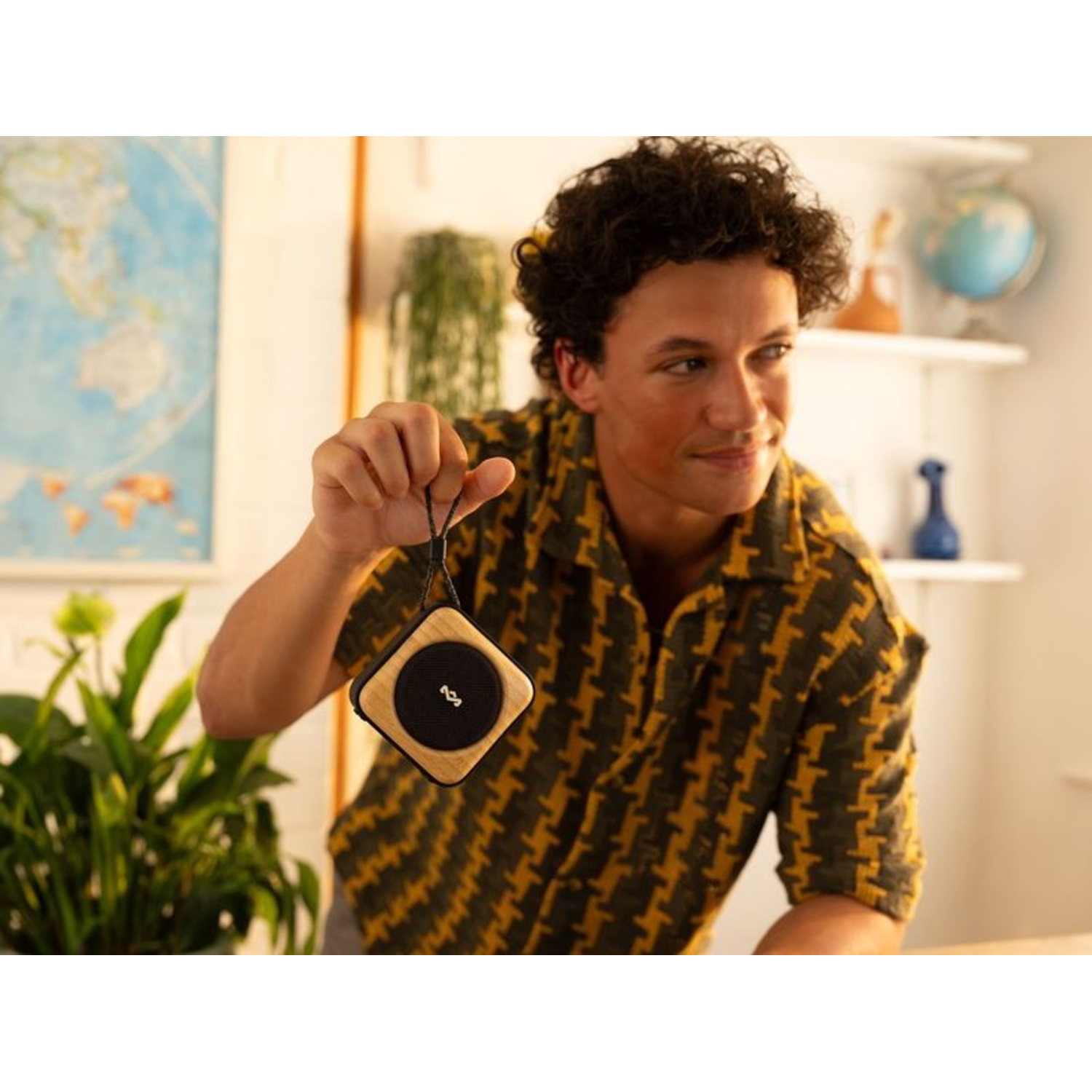 A man holds a small speaker in his hand, smiling, with a map and shelf in the background.