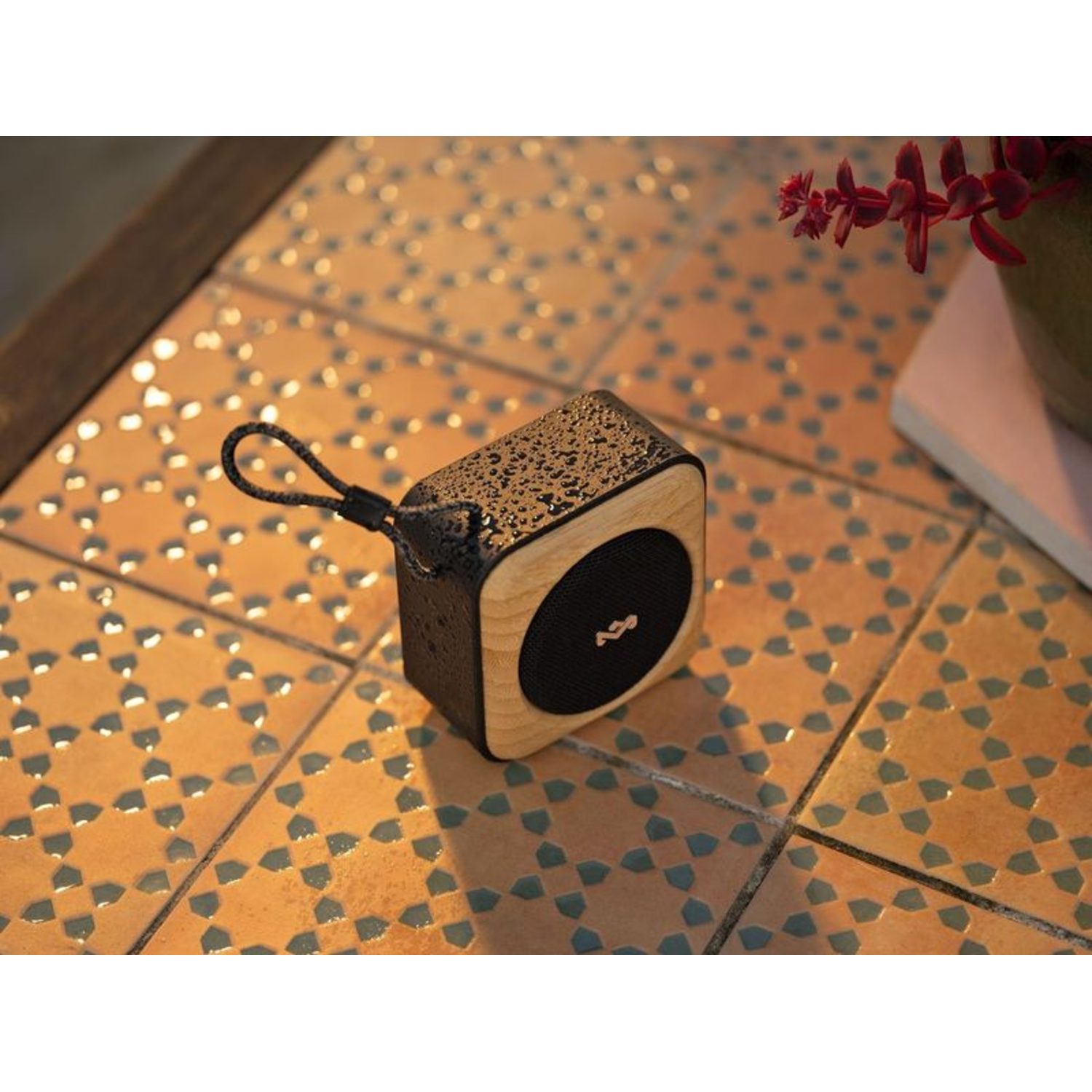 Small, black and wood speaker on a tiled table. Red flowers are in the background.