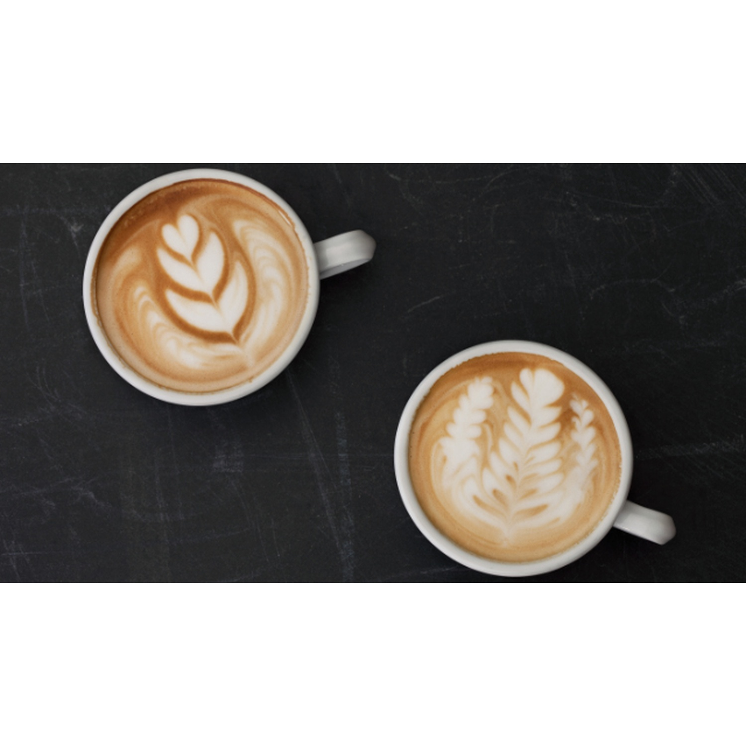 Due tazze di caffè con latte art su una superficie scura, vista dall'alto. Ogni tazza ha un disegno a forma di foglia.
