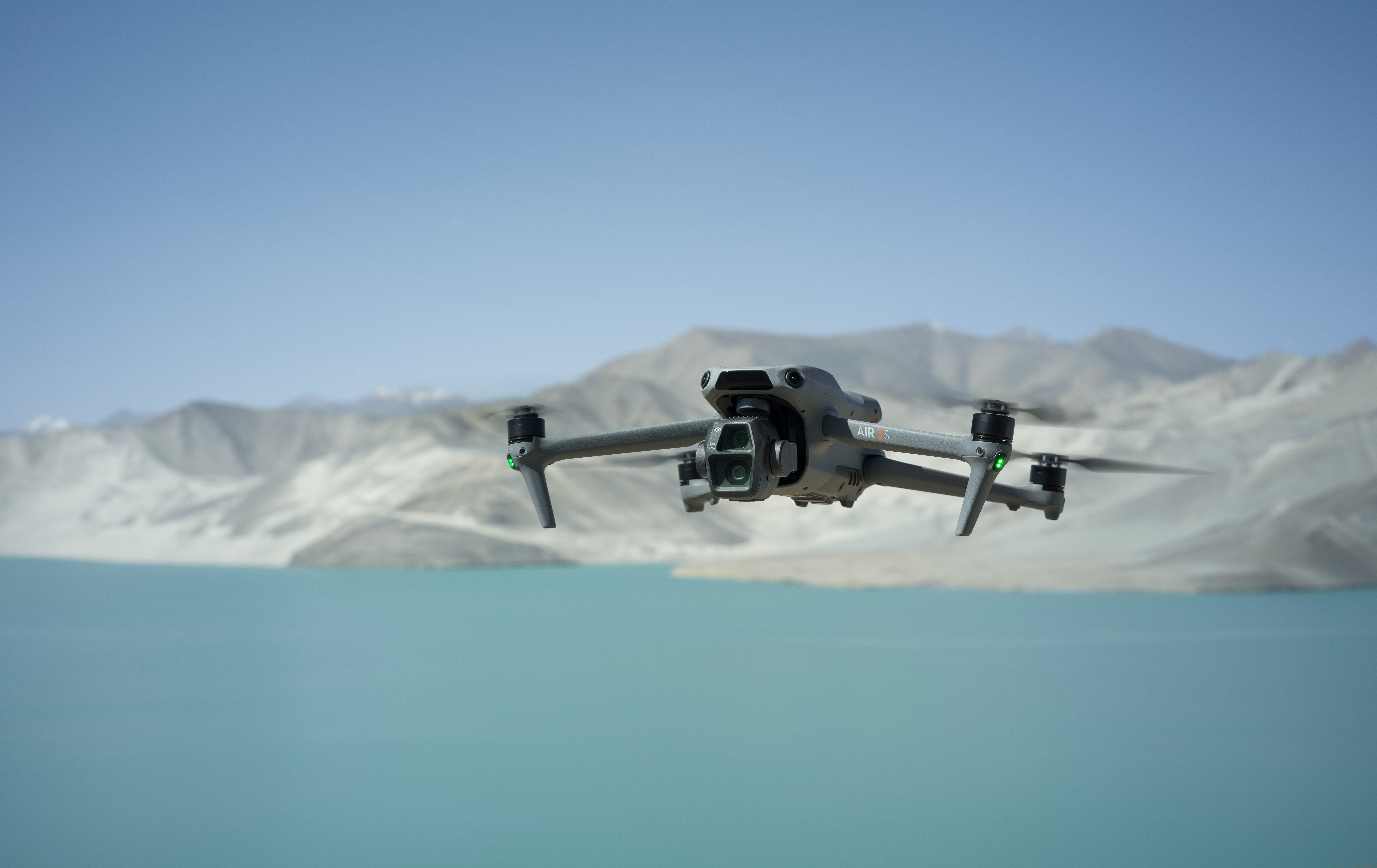 Dron lecący nad wodą i górami pod błękitnym niebem. Dron ma trzy kamery.