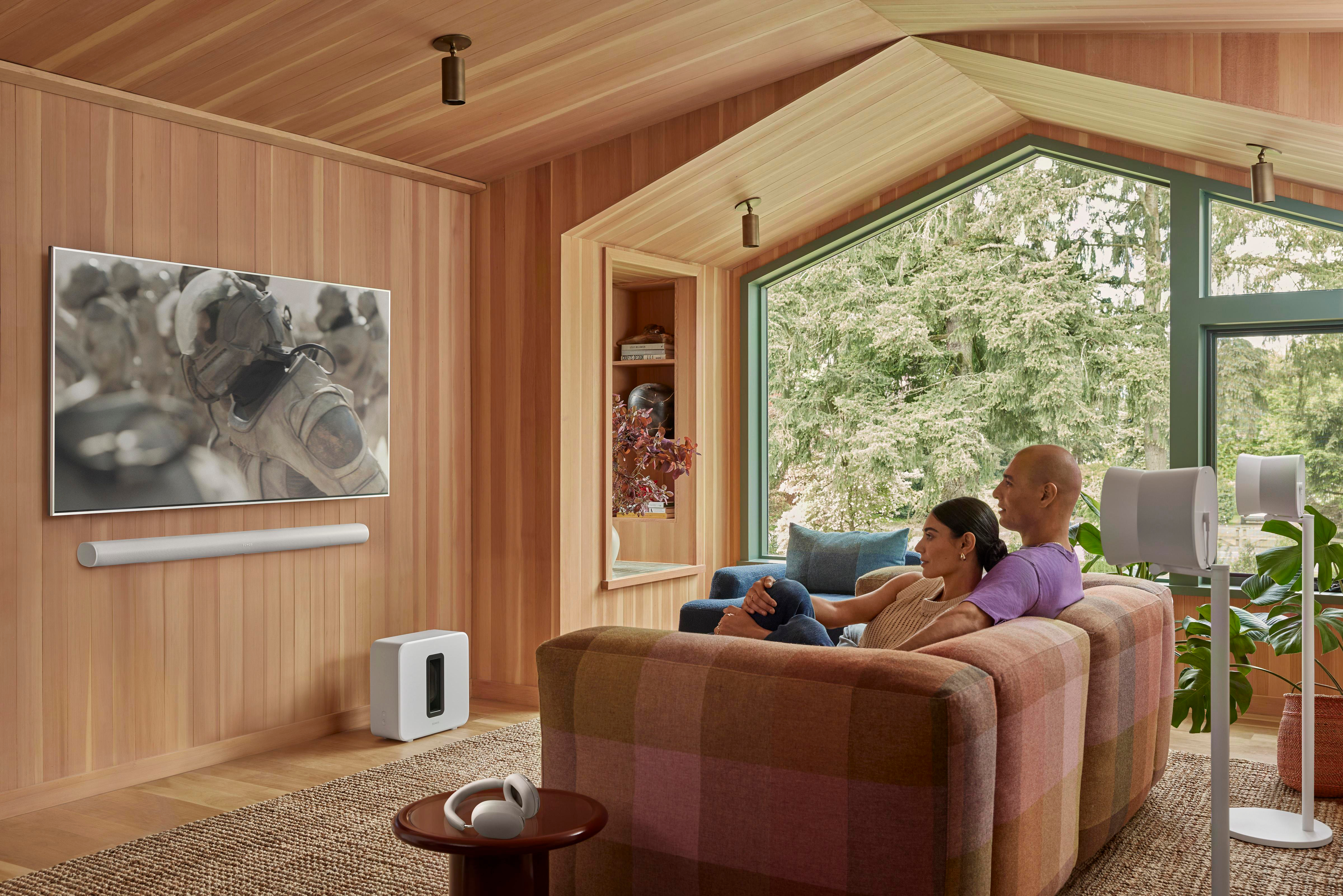 Wohnzimmer mit Paar auf Couch, das fernsieht, Lautsprecher links und rechts.