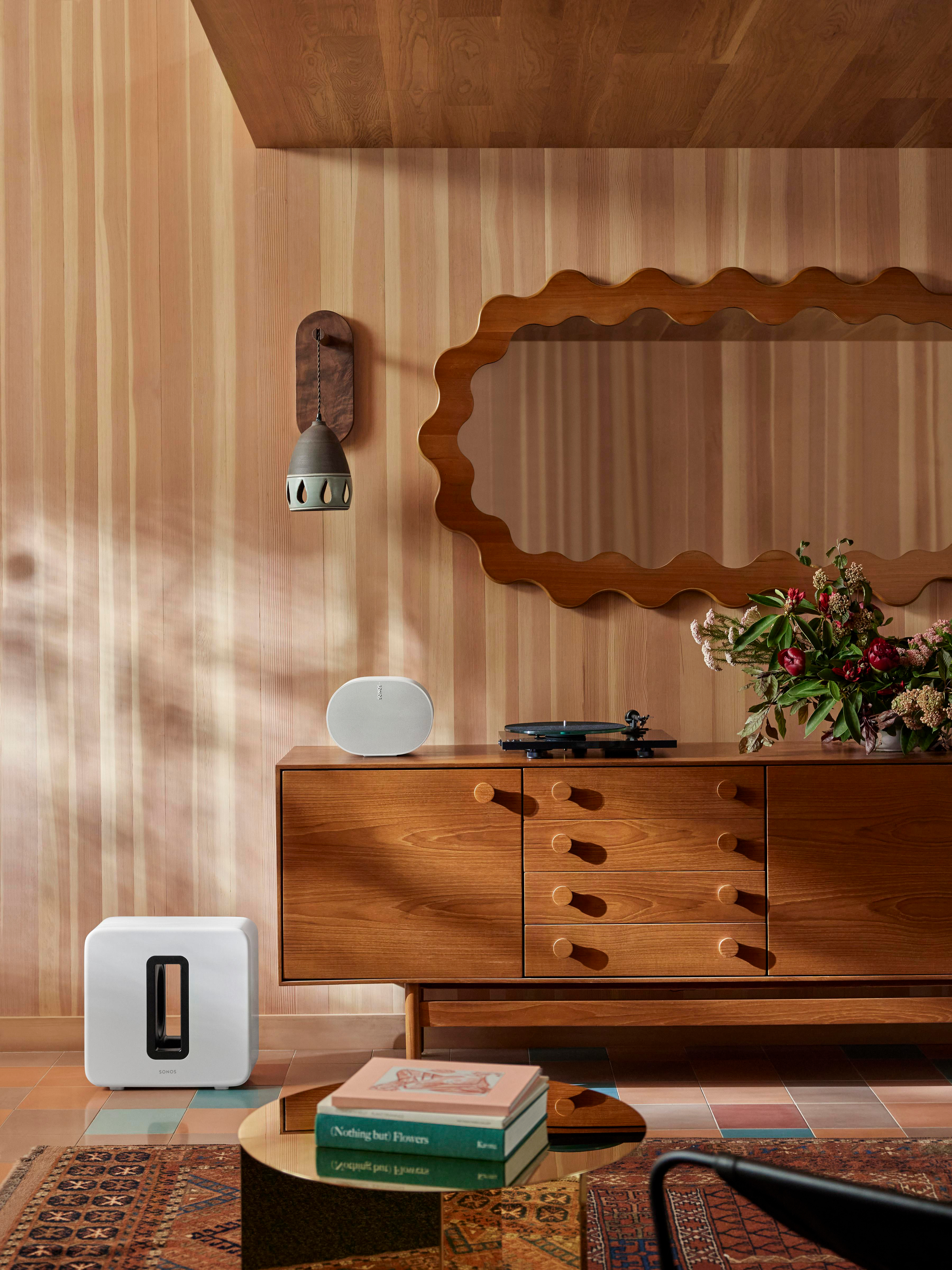 Ein Wohnzimmer mit einem weißen Lautsprecher, einem Plattenspieler und einem Holzschrank. Es gibt einen Spiegel und einen runden Couchtisch im Raum.