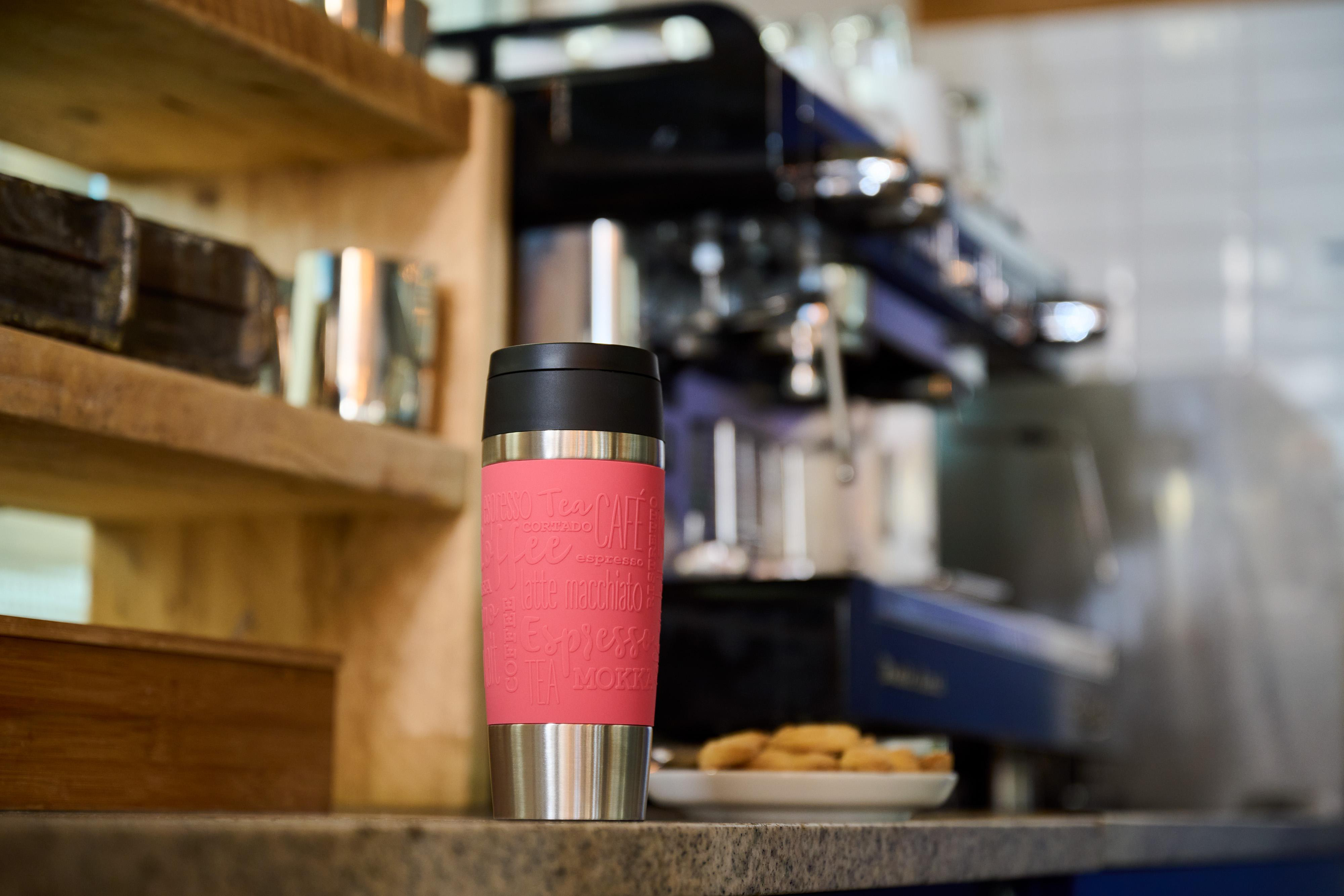 Ein rosa-silberner Reisebecher steht auf einer Theke. Im Hintergrund befindet sich eine Kaffeemaschine.