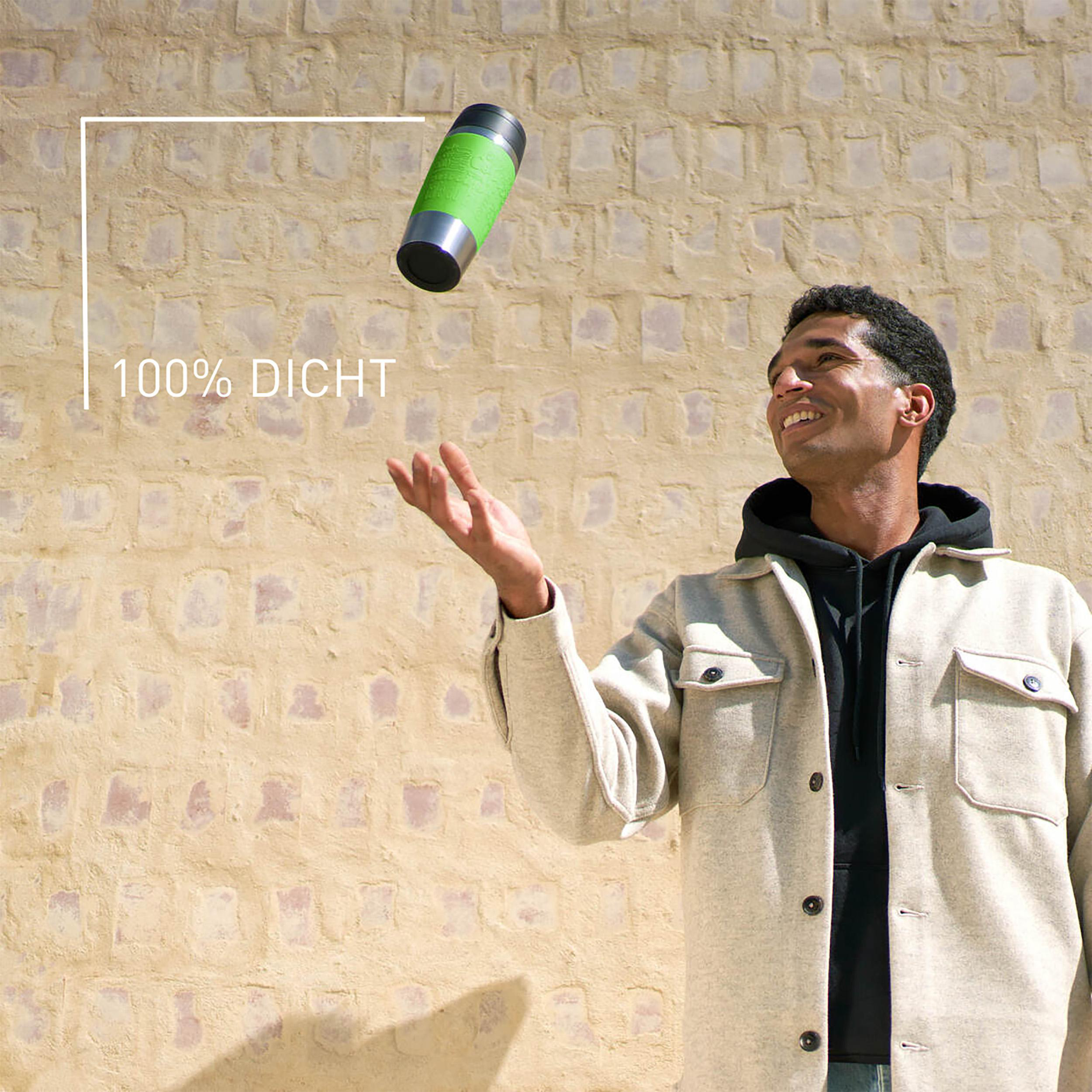 Ein Mann in einer Jacke wirft eine grün-silberne Flasche vor eine beige Wand.