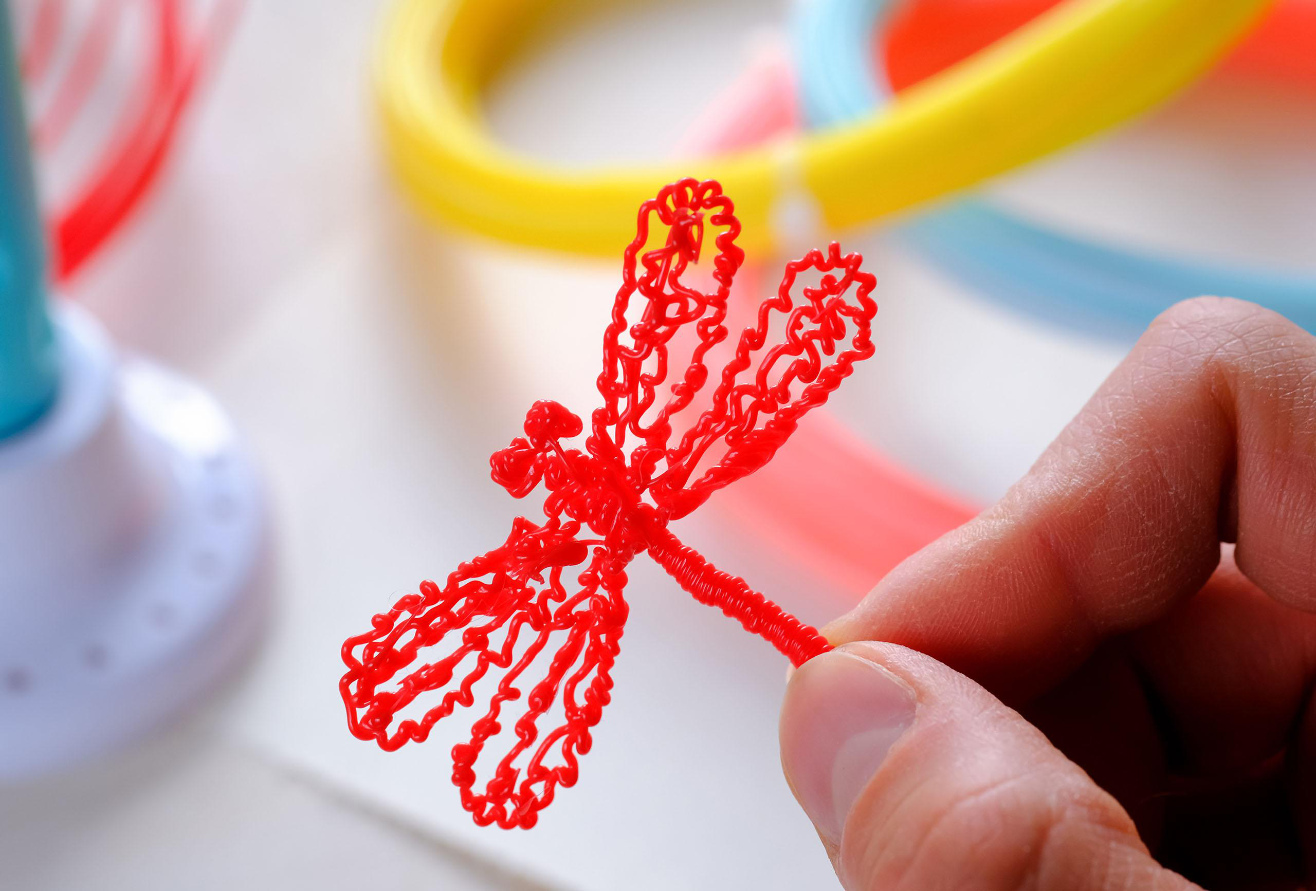 Eine rote Libelle aus Kunststofffilament in einer Hand. Im Hintergrund farbiges Kunststofffilament.