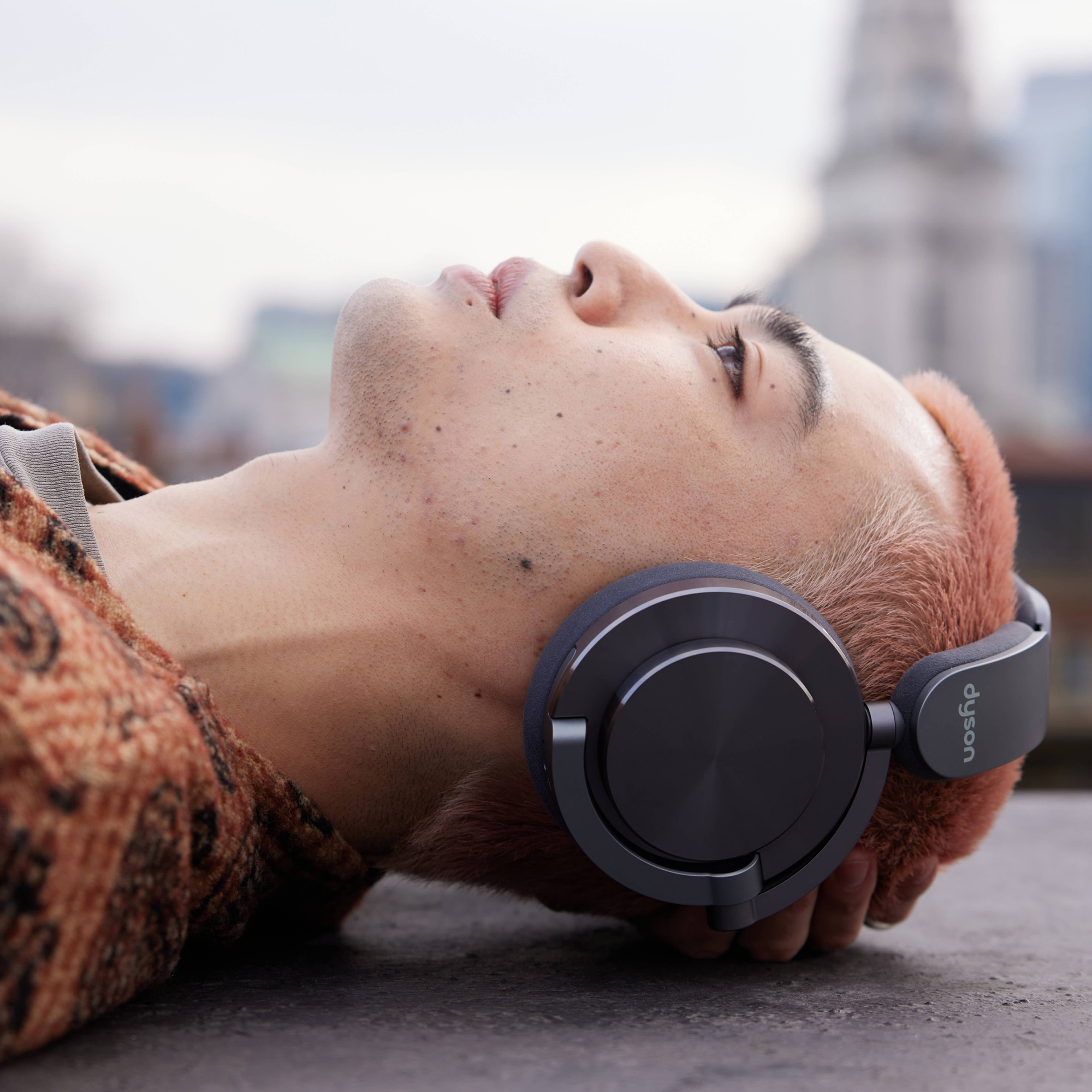 Mężczyzna w słuchawkach Dyson, leżący i patrzący w górę. W tle rozmyte budynki miejskie.