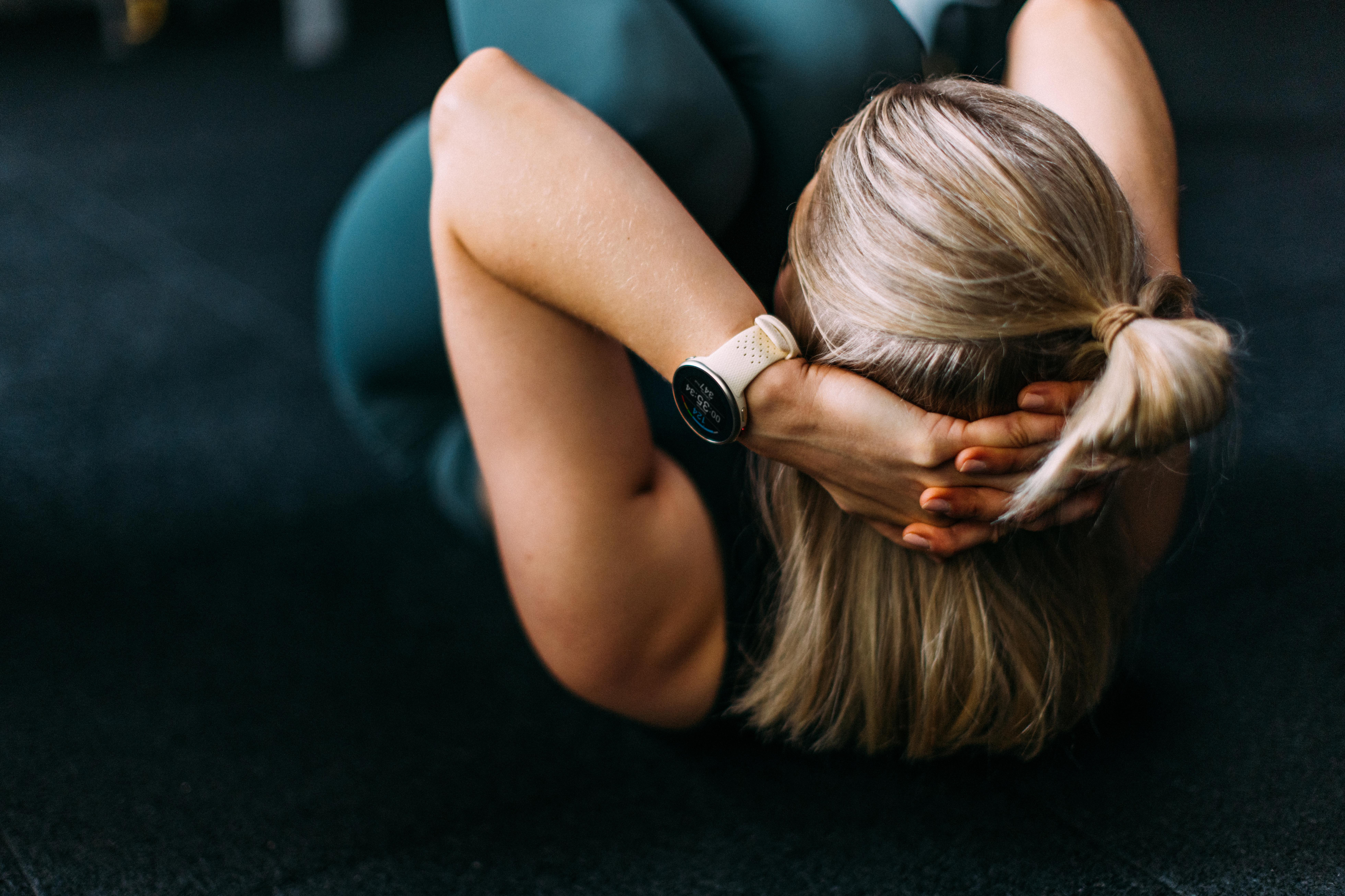 Eine Frau macht Sit-ups, Hände hinter dem Kopf, trägt eine Smartwatch. Fitnessstudio-Umgebung.