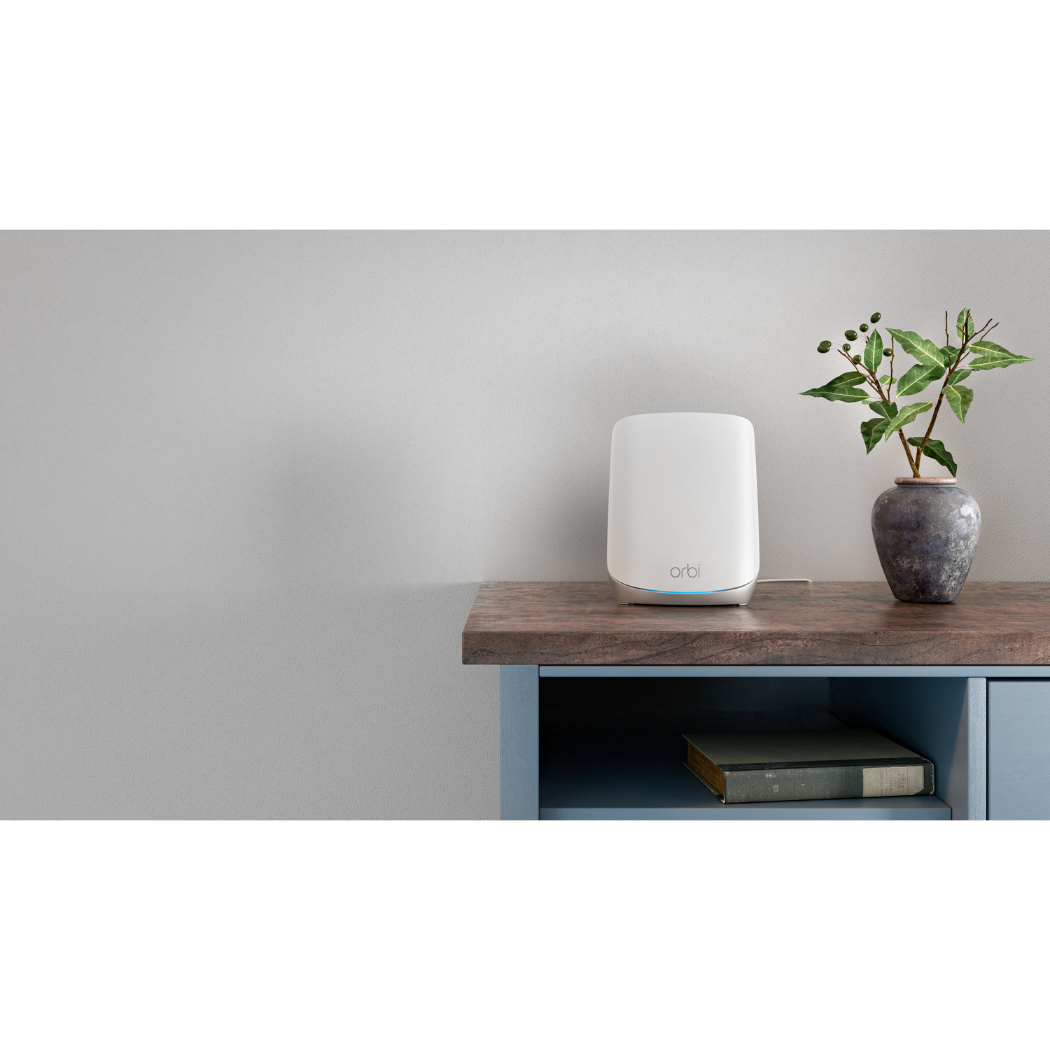 Un routeur blanc sur un meuble bleu. Un vase avec une plante est sur le meuble. Le mur est gris.