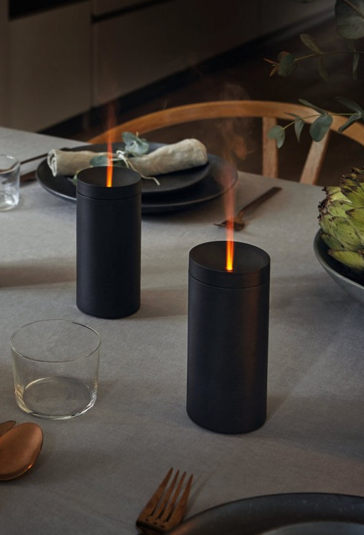 Dwa czarne urządzenia przypominające świece na stole, oświetlone płomieniami. Obok szklanka i widelec.