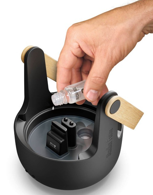 Ręka wlewa płyn z małej butelki do czarnego urządzenia w kształcie jaja z drewnianymi akcentami. Białe tło.