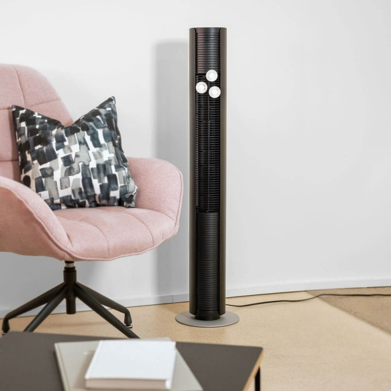 Czarny wysoki wentylator. Stoi na szarej podstawie. Różowy fotel, poduszka i stół w tle.