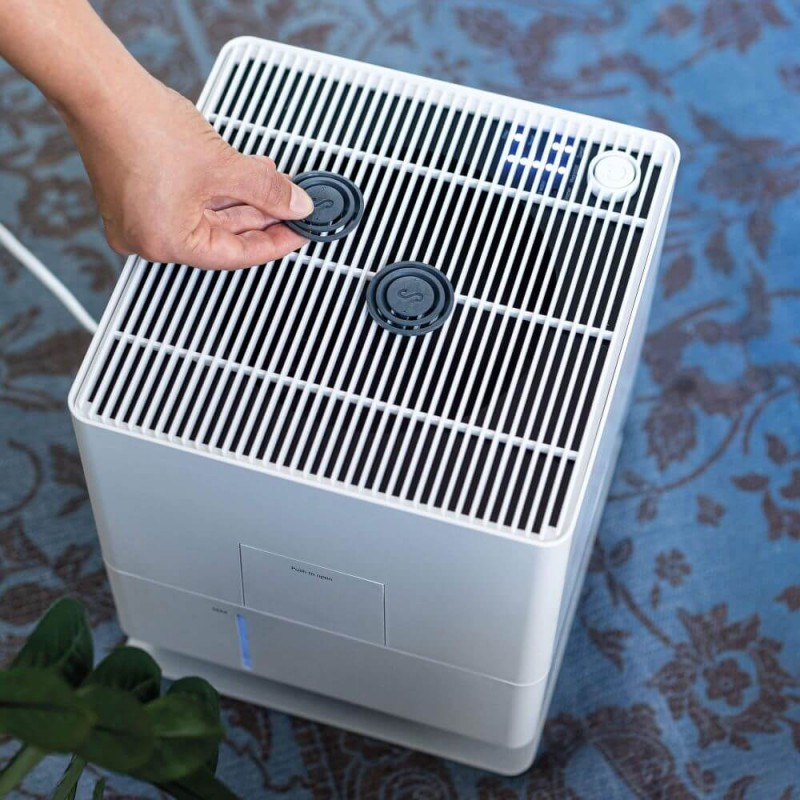 Ręka umieszczająca dwa czarne dyski w białym oczyszczaczu powietrza na niebieskim tle.