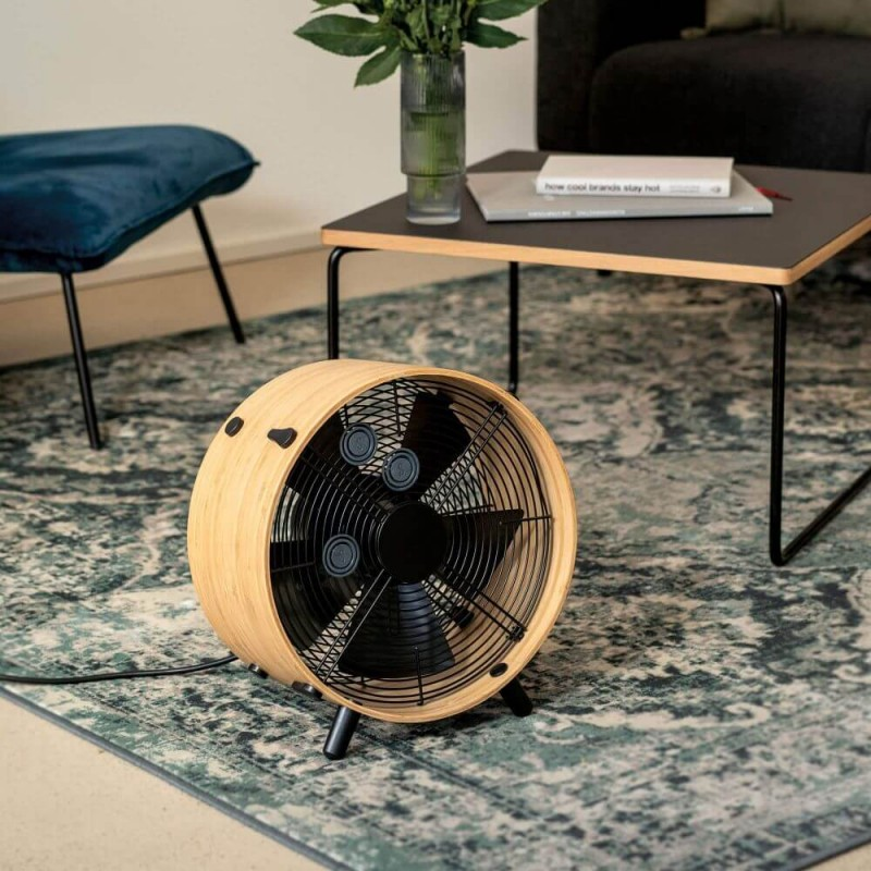 Drewniany wentylator na dywanie. Za nim stół z książką i wazonem. Po lewej niebieski stołek.