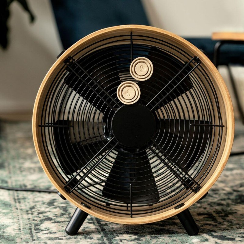 Okrągły wentylator z drewnianym obramowaniem i czarnymi łopatkami. Stoi na wzorzystym dywanie.